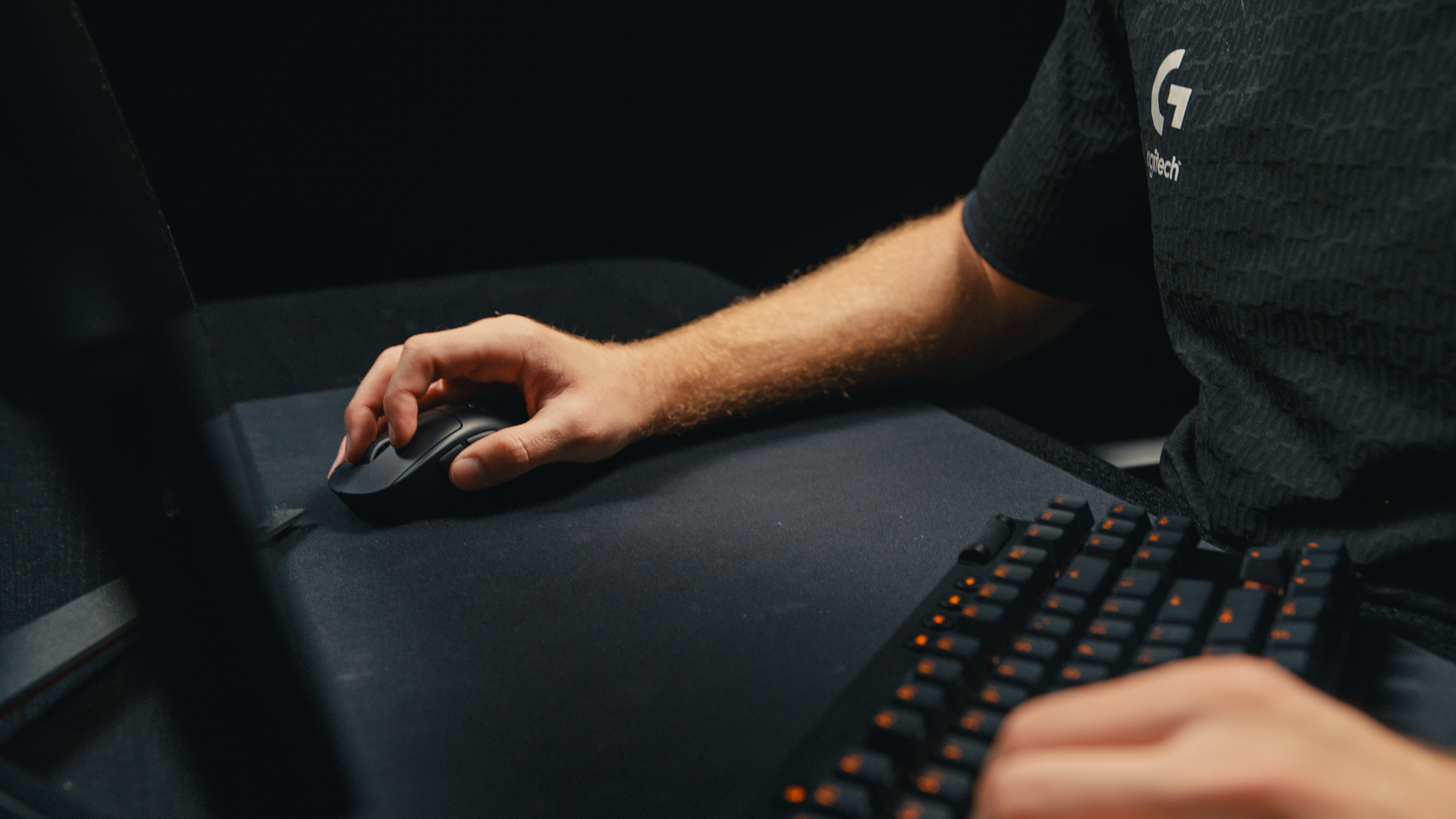Ręka używa czarnej myszy na czarnej podkładce. Klawiatura jest na pierwszym planie. Widoczna jest czarna koszulka z logo.
