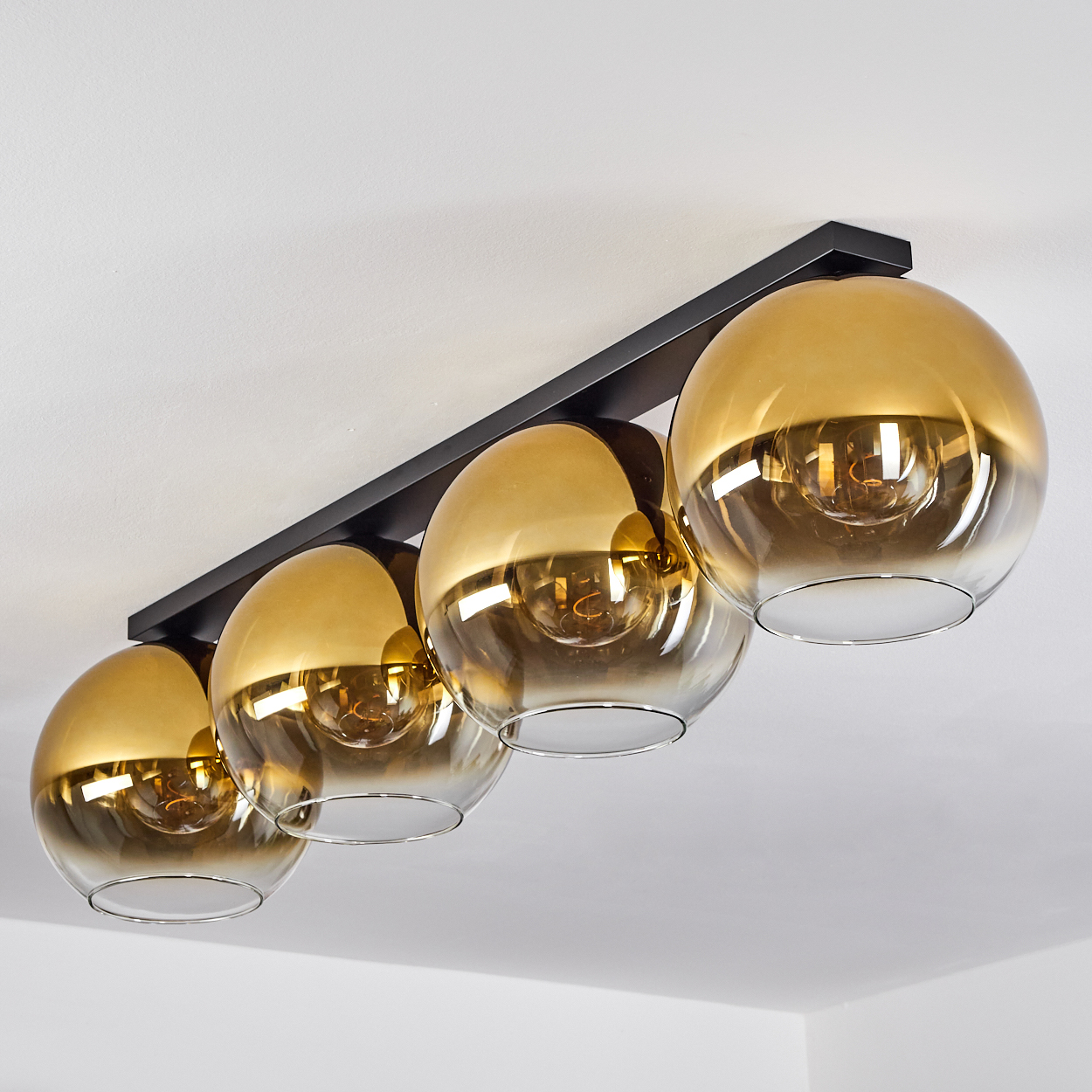 Eine Deckenleuchte mit vier goldfarbenen Glaskugeln ist an einem schwarzen rechteckigen Rahmen befestigt. Die Decke ist weiß.