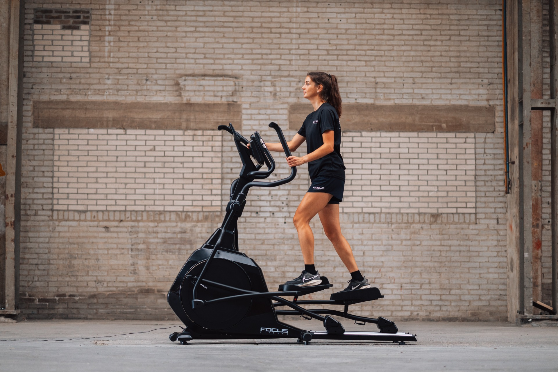 Woman in black activewear exercises on a black elliptical machine. Background is a brick wall.