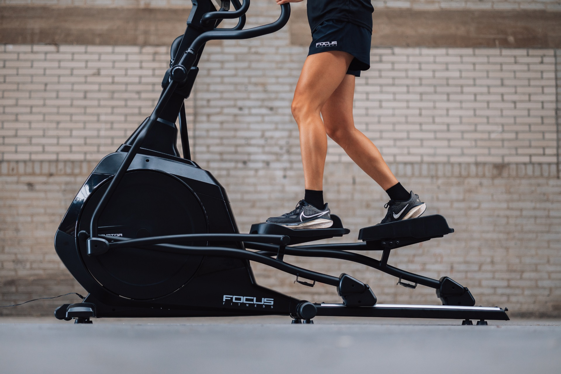 A person is exercising on a black elliptical machine. They are wearing black shorts and sneakers. The background is a brick wall.