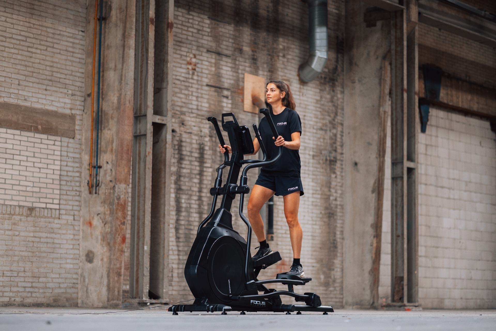 A woman exercises on an elliptical machine in an industrial setting. She wears black athletic clothes. The background is brick.