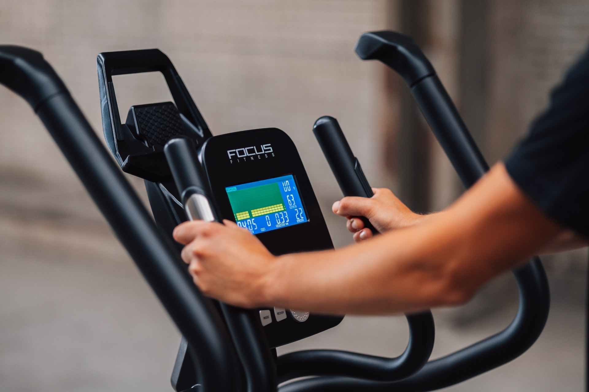 Close-up of exercise machine. Hands on handles, monitor showing stats. Black frame. Background is a blurred wall.