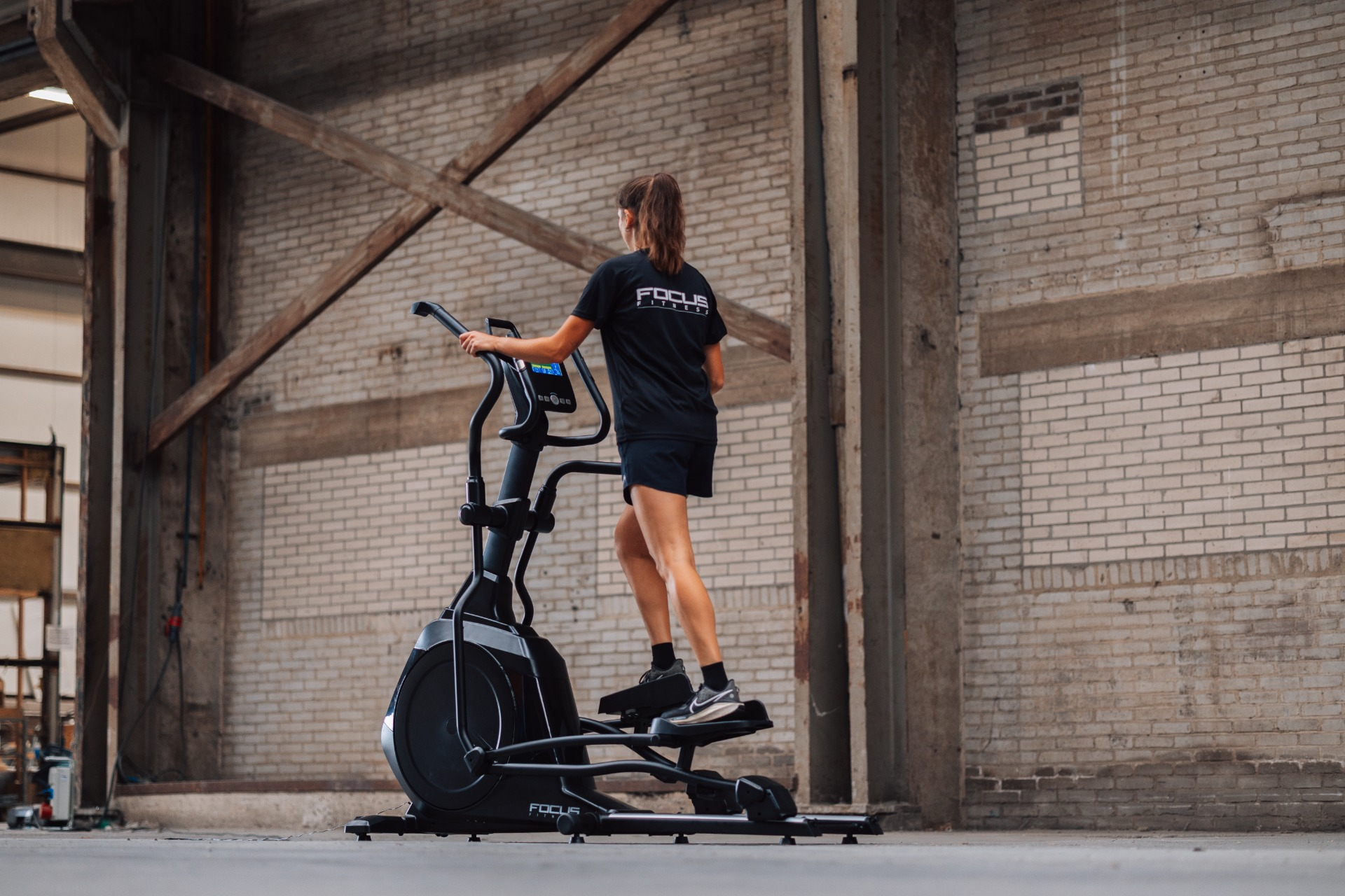 A woman exercises on a black elliptical machine in a brick-walled warehouse. She wears black athletic wear.