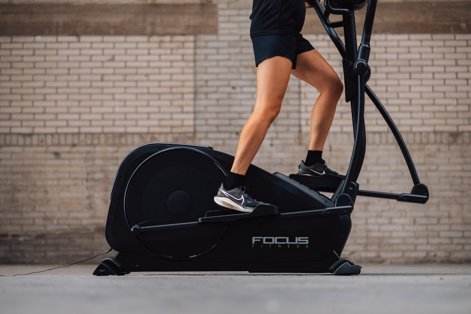 Personne faisant de l'exercice sur un elliptique noir. Le fond est un mur de briques. La machine est étiquetée FOCUS FITNESS.