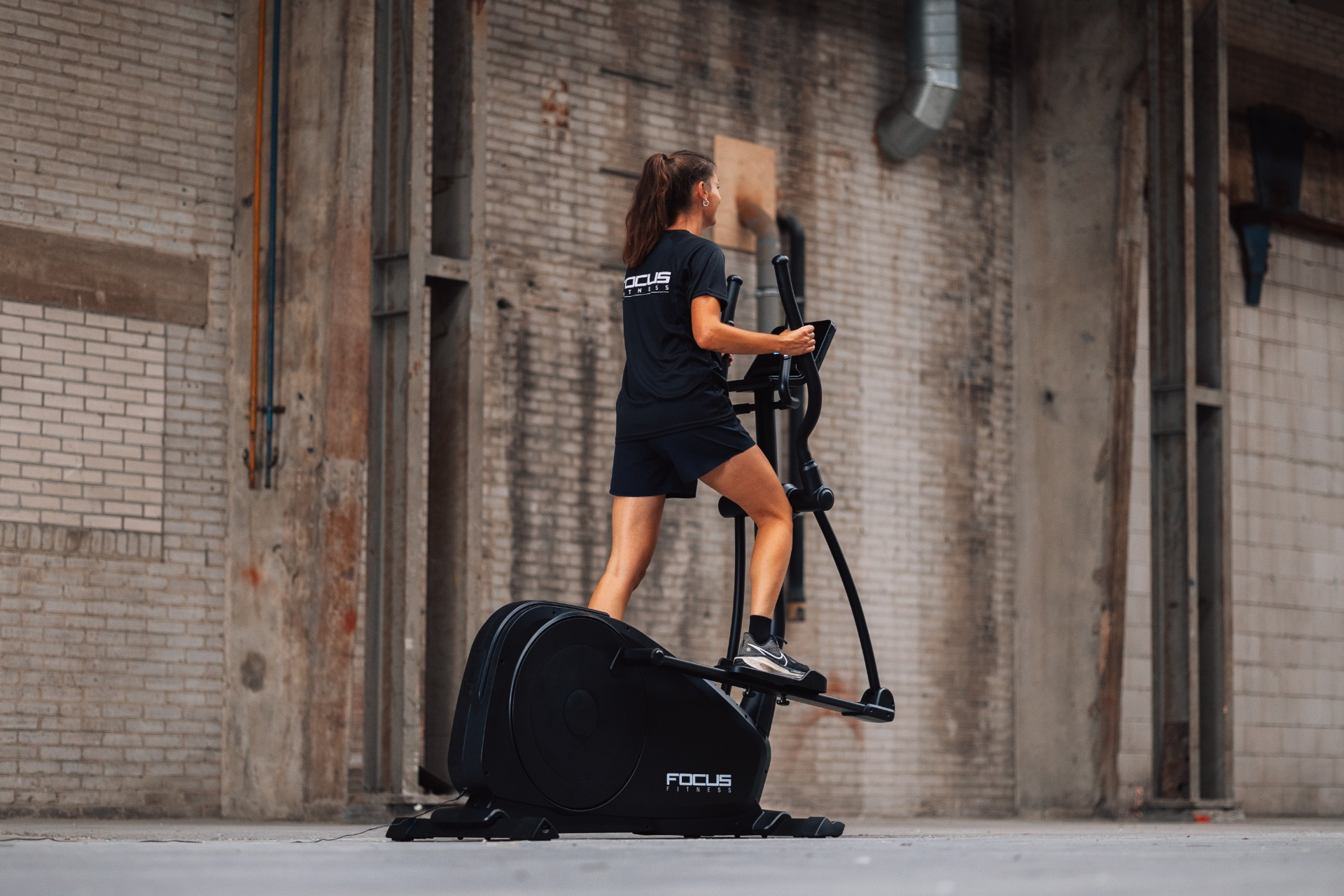 Une femme en short et t-shirt noirs s'exerce sur un elliptique noir à l'intérieur d'un bâtiment aux murs de briques.