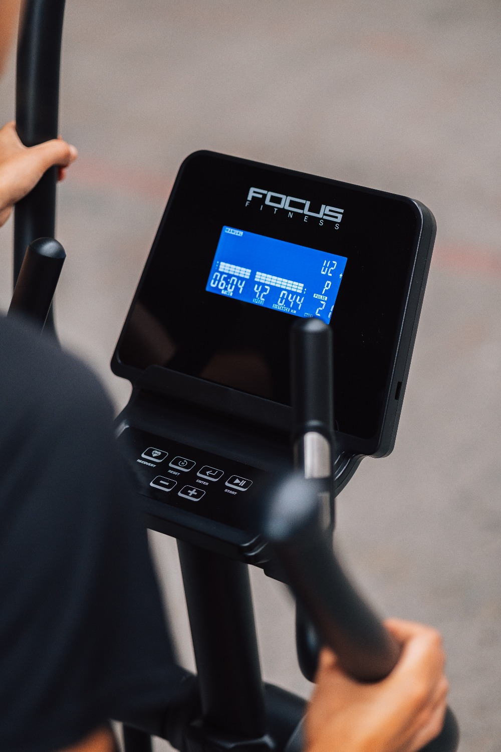 Personne utilisant un vélo elliptique Focus Fitness. Affiche la vitesse, la distance et d'autres mesures sur écran bleu. Poignées noires.