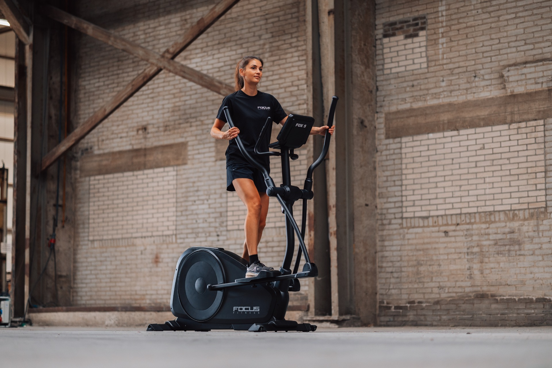 Une femme s'entraîne sur un elliptique dans un entrepôt aux murs de briques. Elle porte des vêtements d'entraînement noirs. Focus Fitness FOX 5 est visible.