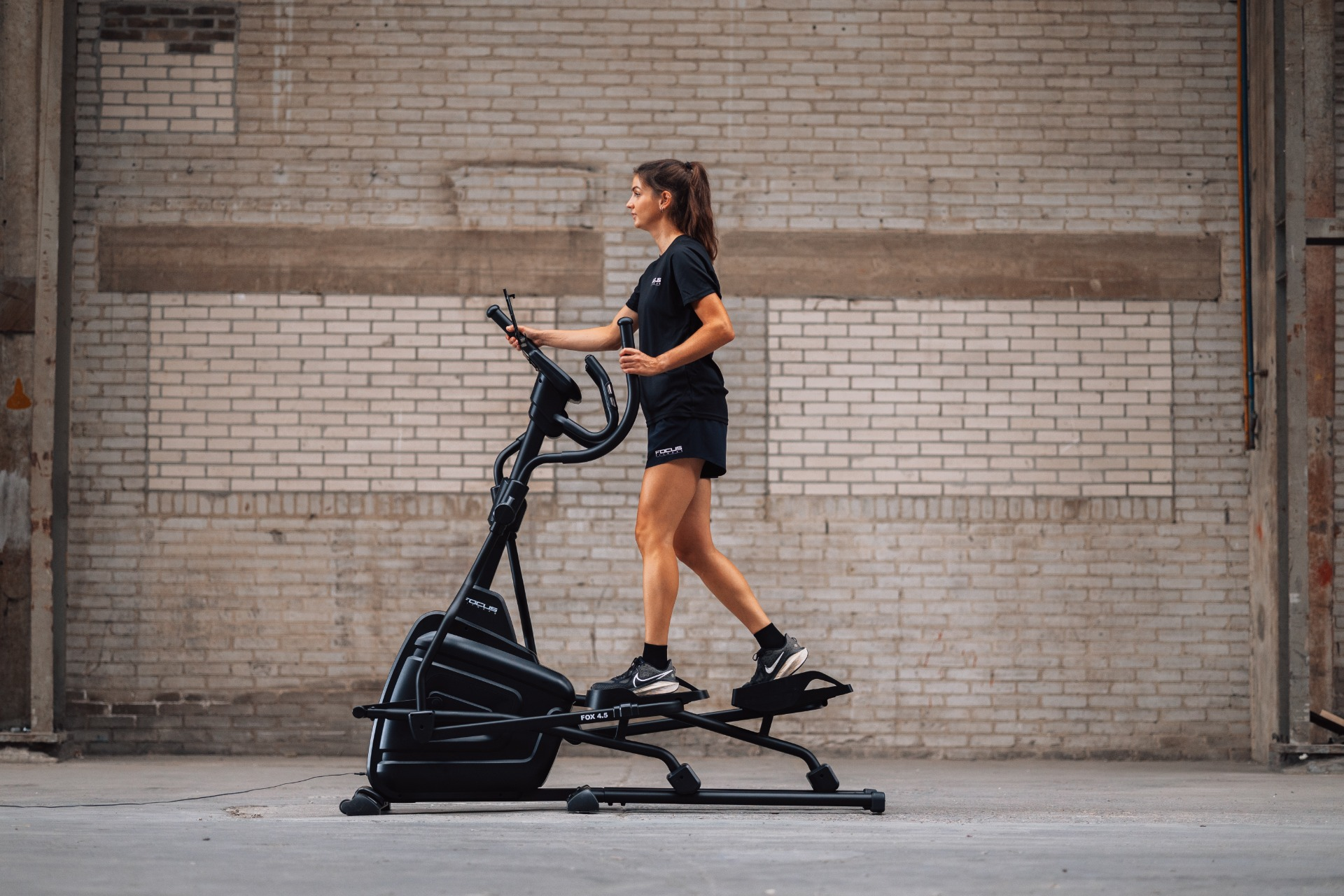 Woman in black activewear on elliptical machine. Brick wall background. Focus 4.5 model. Exercising indoors.