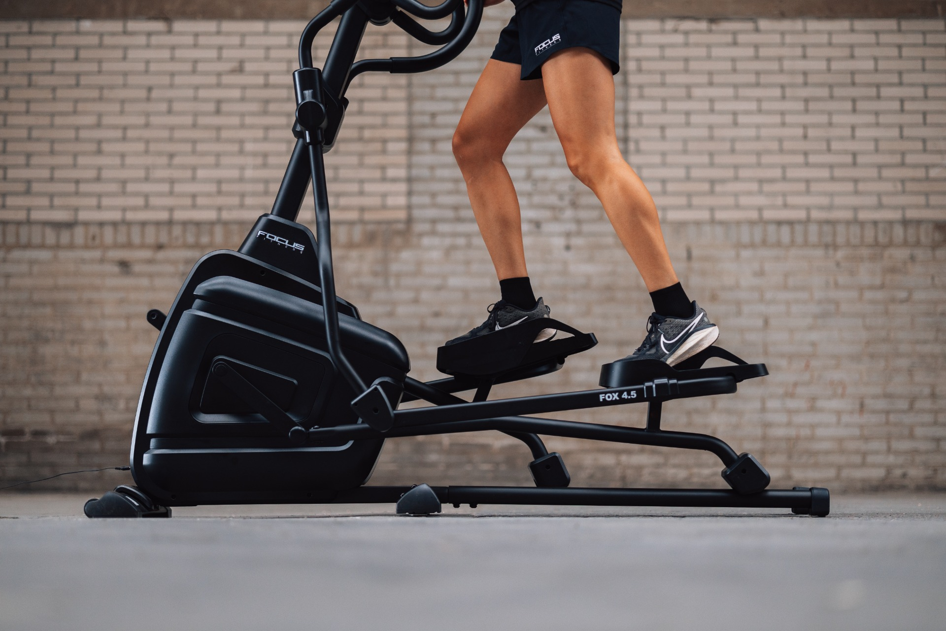 A person is using a black elliptical machine. The background is a brick wall. They are wearing shorts and sneakers.