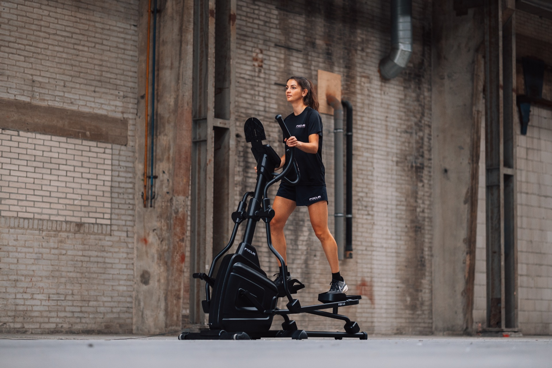 A woman exercises on an elliptical machine in front of a brick wall. She wears black sportswear and sneakers.