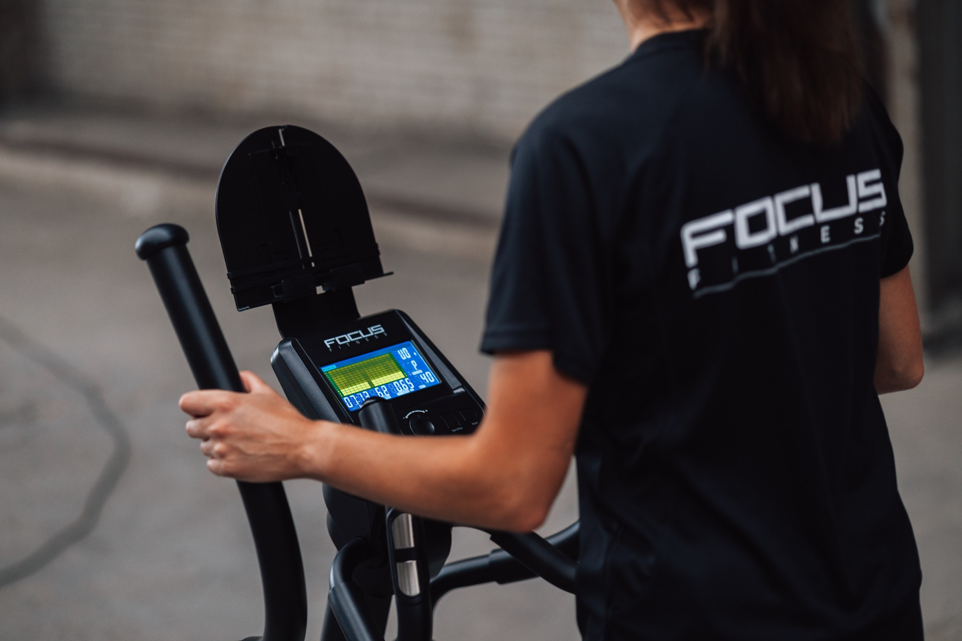 A person wearing a black Focus Fitness shirt uses an elliptical machine. The display shows workout data.