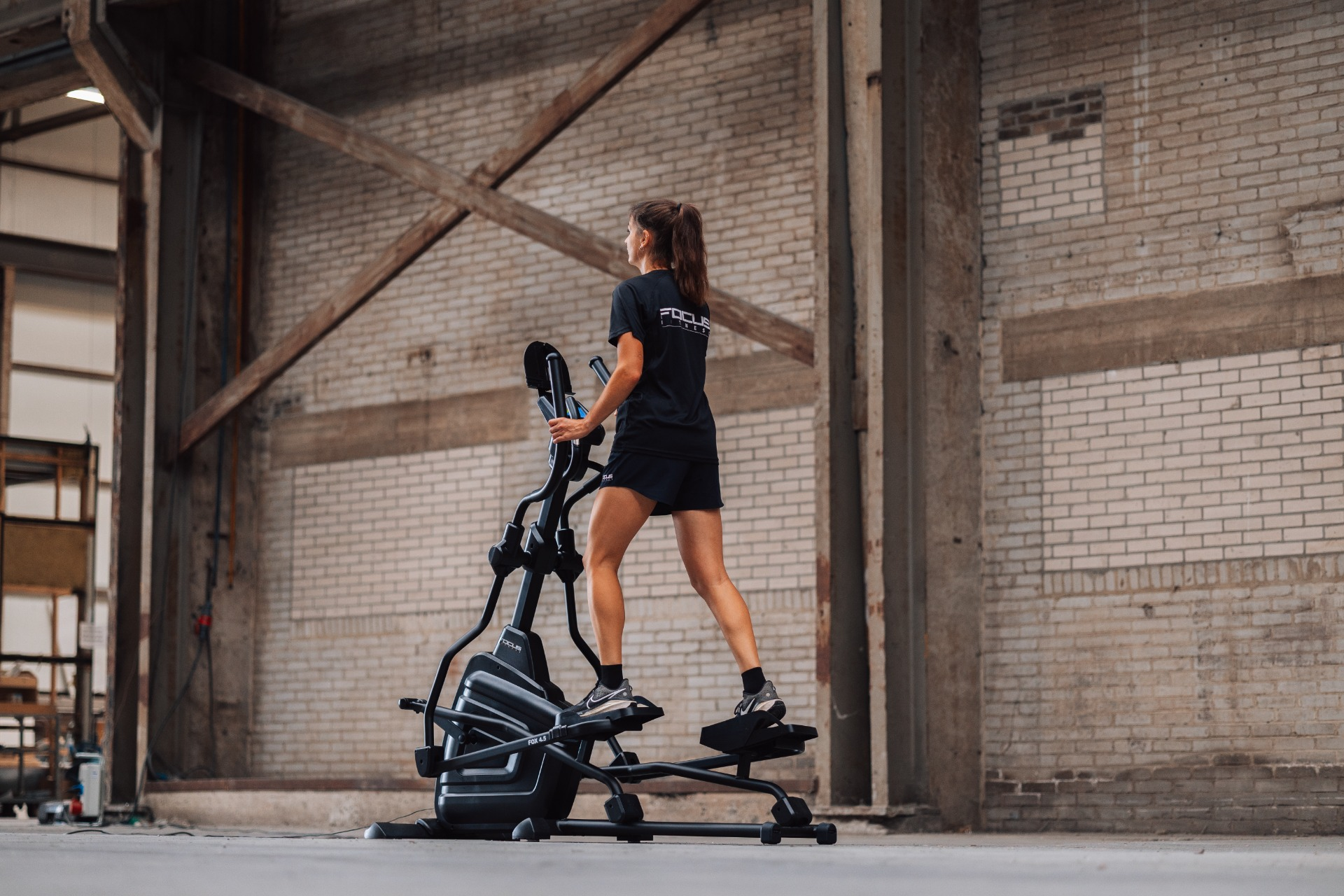 A woman exercises on an elliptical trainer in a brick-walled industrial space. She wears black shorts and a shirt.