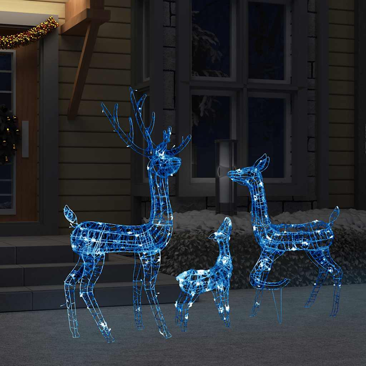 Drei beleuchtete blaue Rentierdekorationen stehen auf einer Veranda. Im Hintergrund ein Haus mit dekorierter Tür und schneebedeckten Büschen.