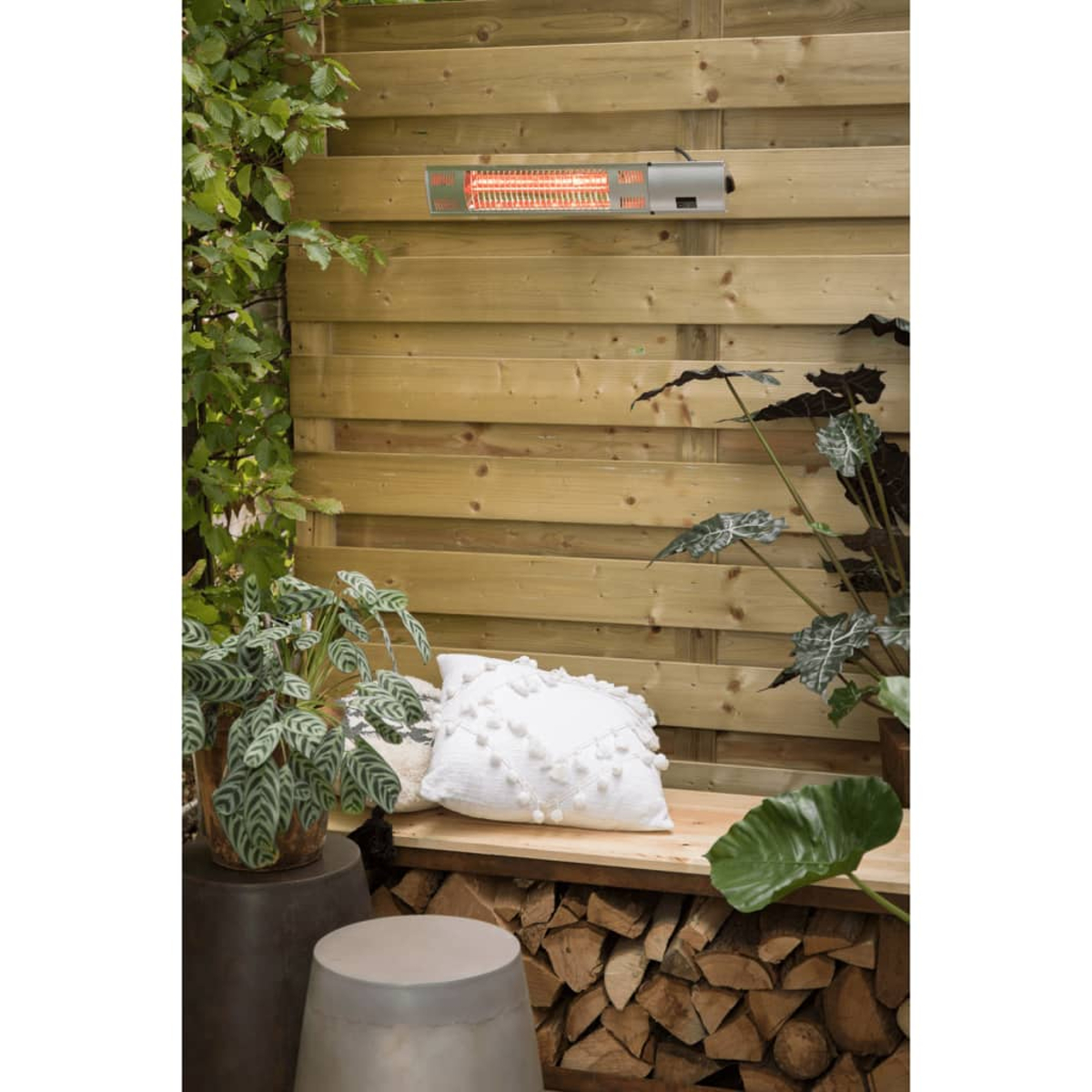 Outdoor scene with wooden fence, heater, bench with white pillow. Plants and logs are also present in the scene.