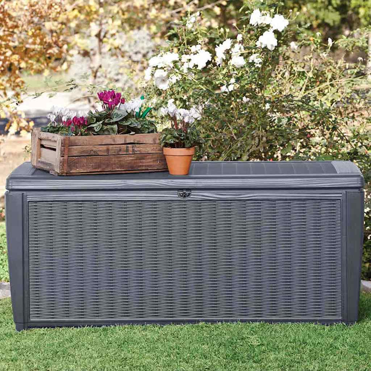 Graue Aufbewahrungsbox auf Gras mit Holzkiste und Topf mit Blumen. Büsche im Hintergrund.