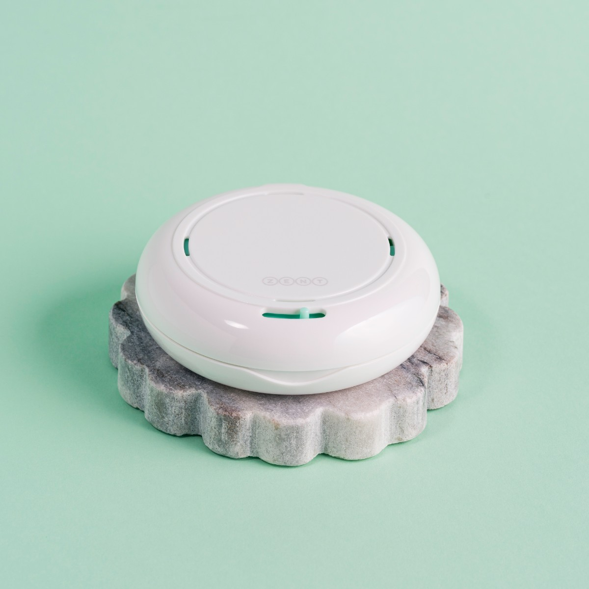 Białe, okrągłe urządzenie z napisem 'ZENT' na górze, umieszczone na szarej, dekoracyjnej kamiennej podstawie, na jasnym zielonym tle.