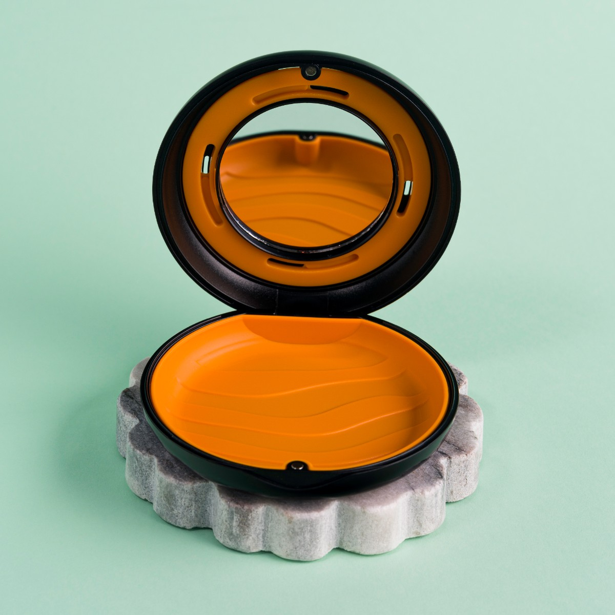 Otwarty, czarno-pomarańczowy kompakt na szarej kamieniu, na miętowym tle. Kompakt ma lusterko i pomarańczową podstawę.