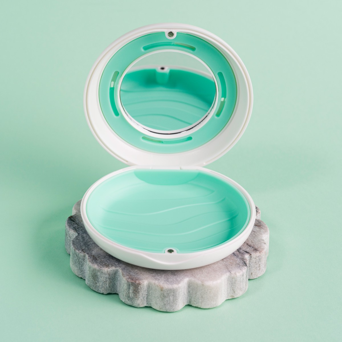 Otwarty biały i turkusowy kompakt na kamiennej podstawie, na jasnym zielonym tle. Lustro wewnątrz pokrywy. Kompakt jest otwarty.