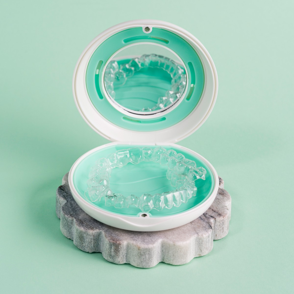 Przezroczyste nakładki na zęby w etui, na marmurowej podstawie, na miętowym tle. Etui jest otwarte, pokazując nakładki.