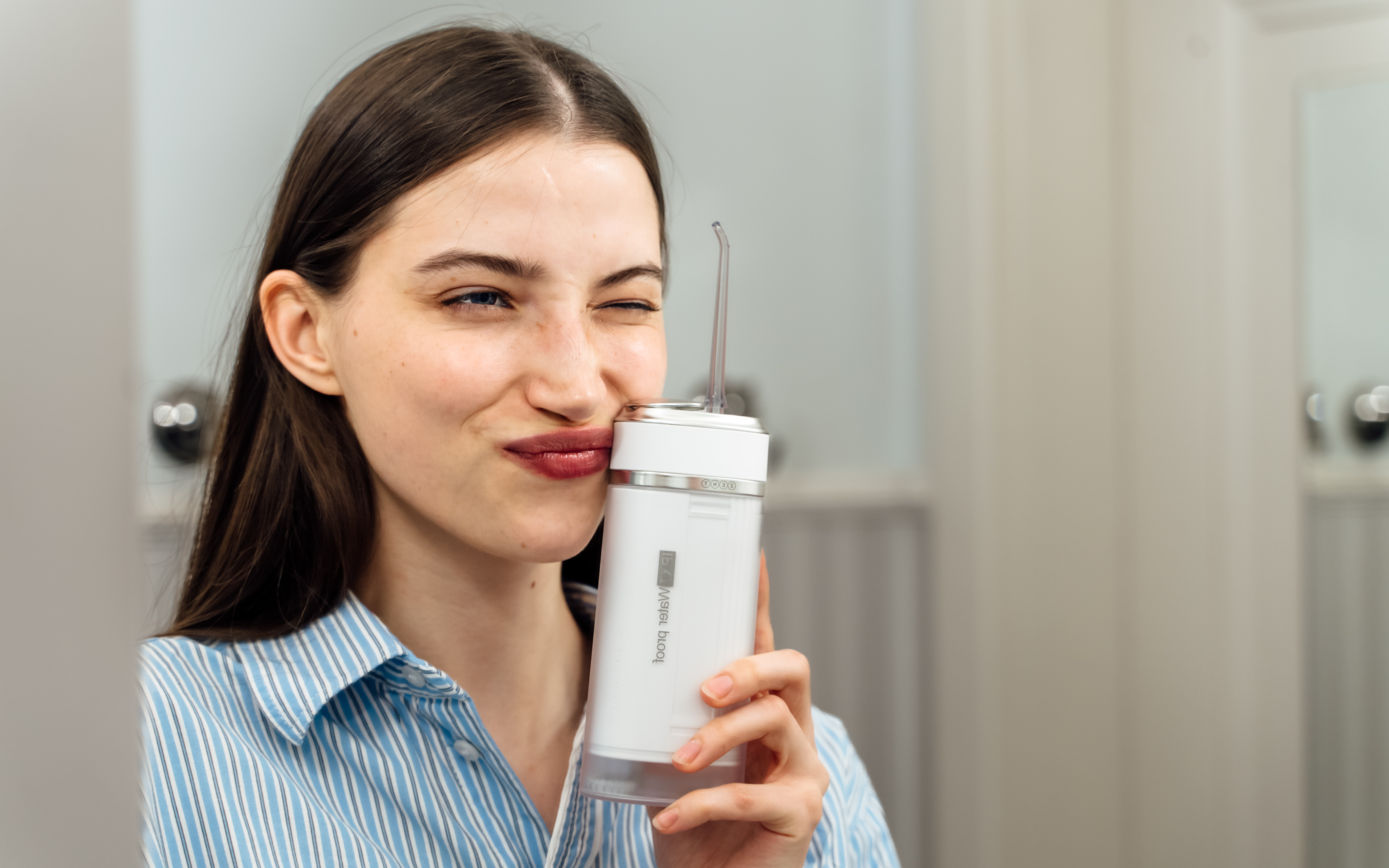 Kobieta trzymająca biały irygator do zębów, mrugająca. Ma na sobie niebieską koszulę w paski. Rozmyte tło łazienki.