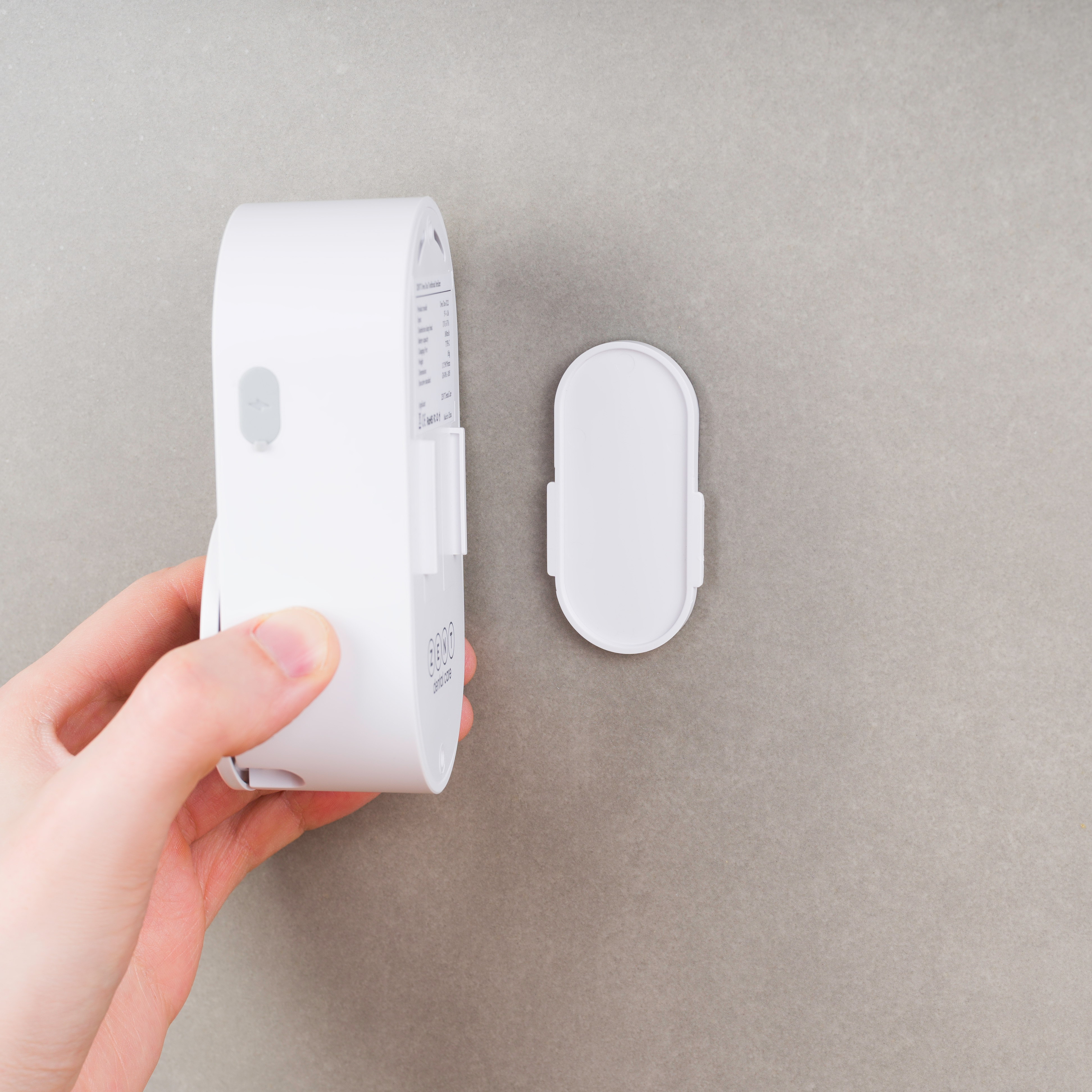 Dłoń trzymająca białe urządzenie z oddzielnym elementem na szarym tle. Etykieta widoczna na urządzeniu.
