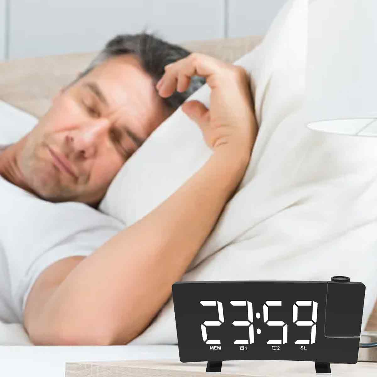 Mann schläft im Bett mit einer Digitaluhr, die 23:59 anzeigt. Die Uhr ist im Vordergrund, Mann im Hintergrund.