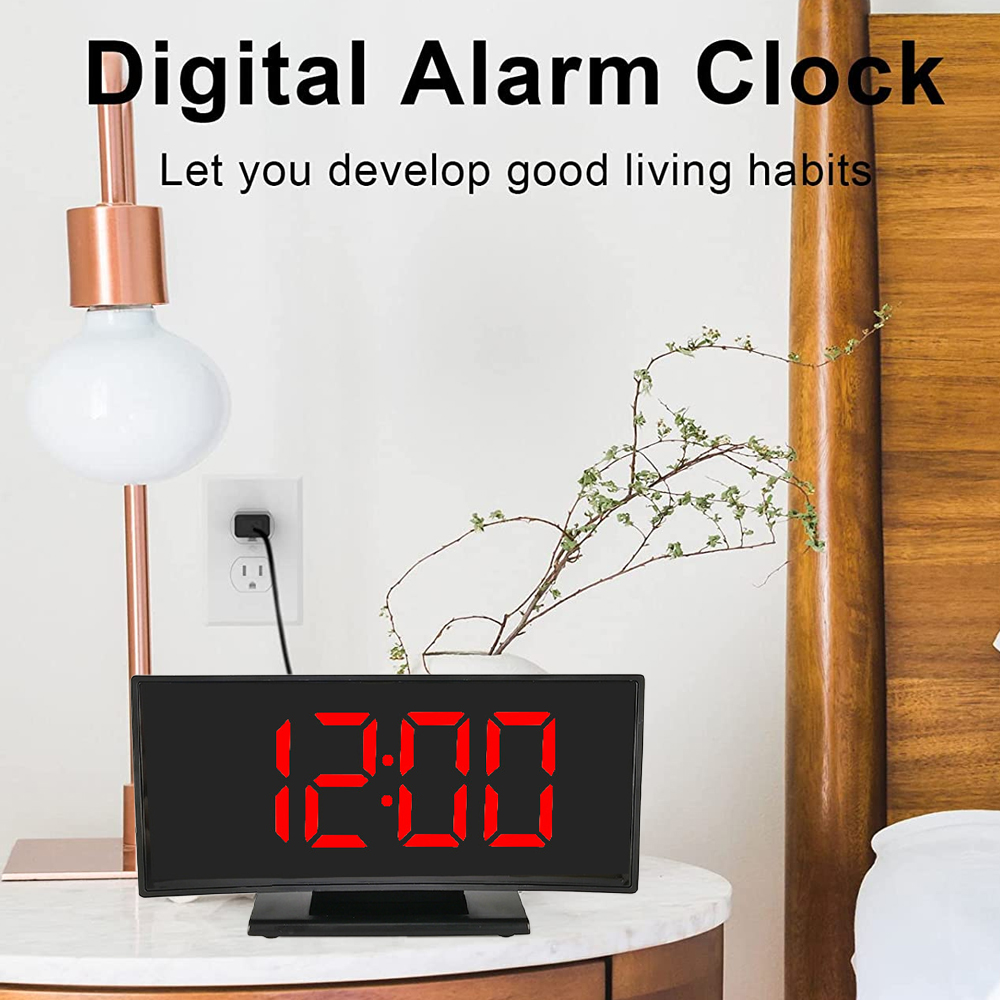 Ein digitaler Wecker mit der Anzeige 12:00 steht auf einem Nachttisch. Eine Lampe und eine Pflanze stehen ebenfalls auf dem Tisch, und ein Bett ist im Hintergrund.