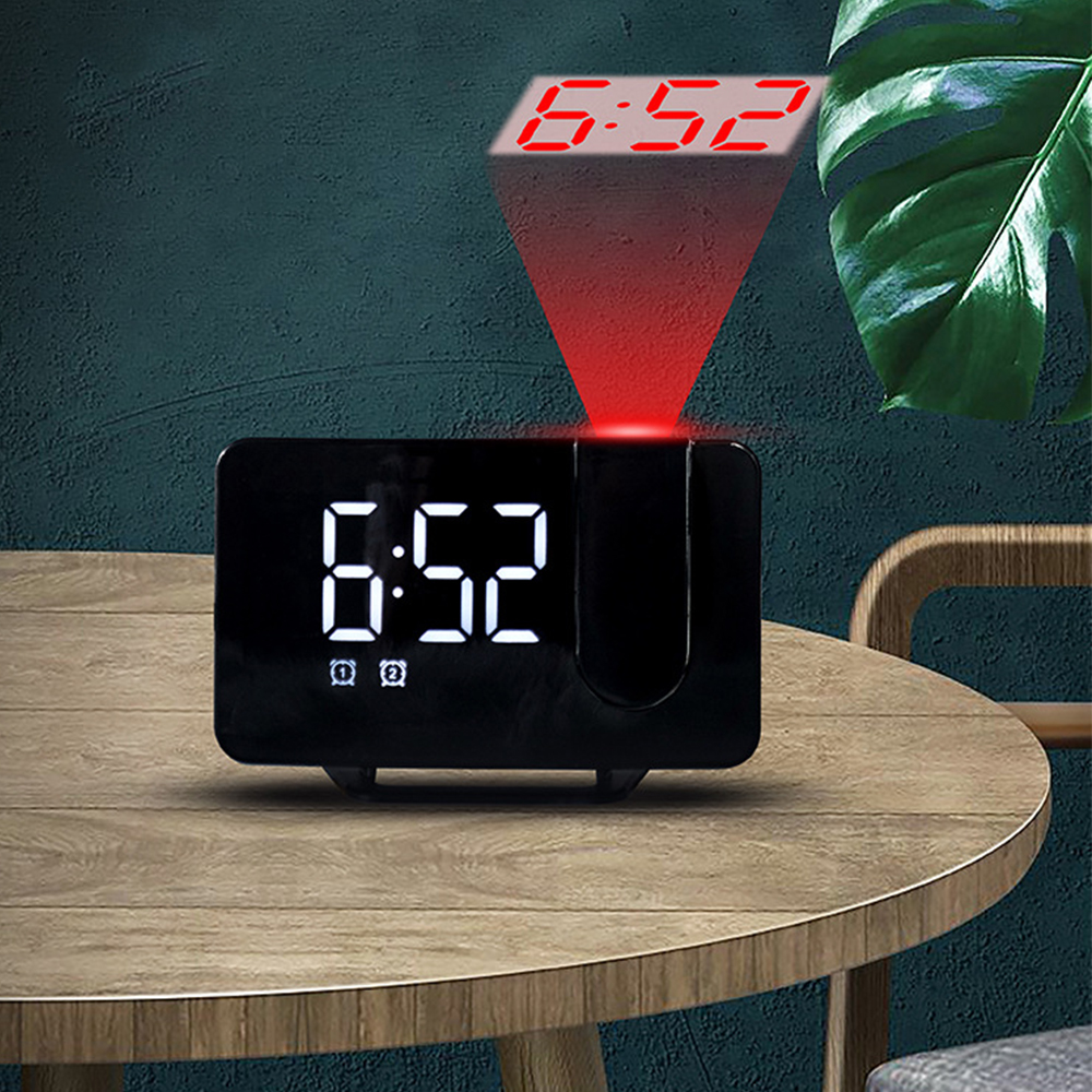 Eine schwarze Digitaluhr zeigt 6:52 an. Sie projiziert die Zeit in Rot an die Wand. Die Uhr steht auf einem Holztisch.