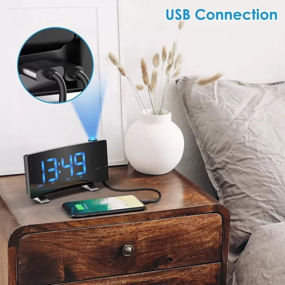 Eine Digitaluhr zeigt 13:49 an. Ein Telefon wird geladen. Eine Vase mit Pflanzen steht neben einem Bett.