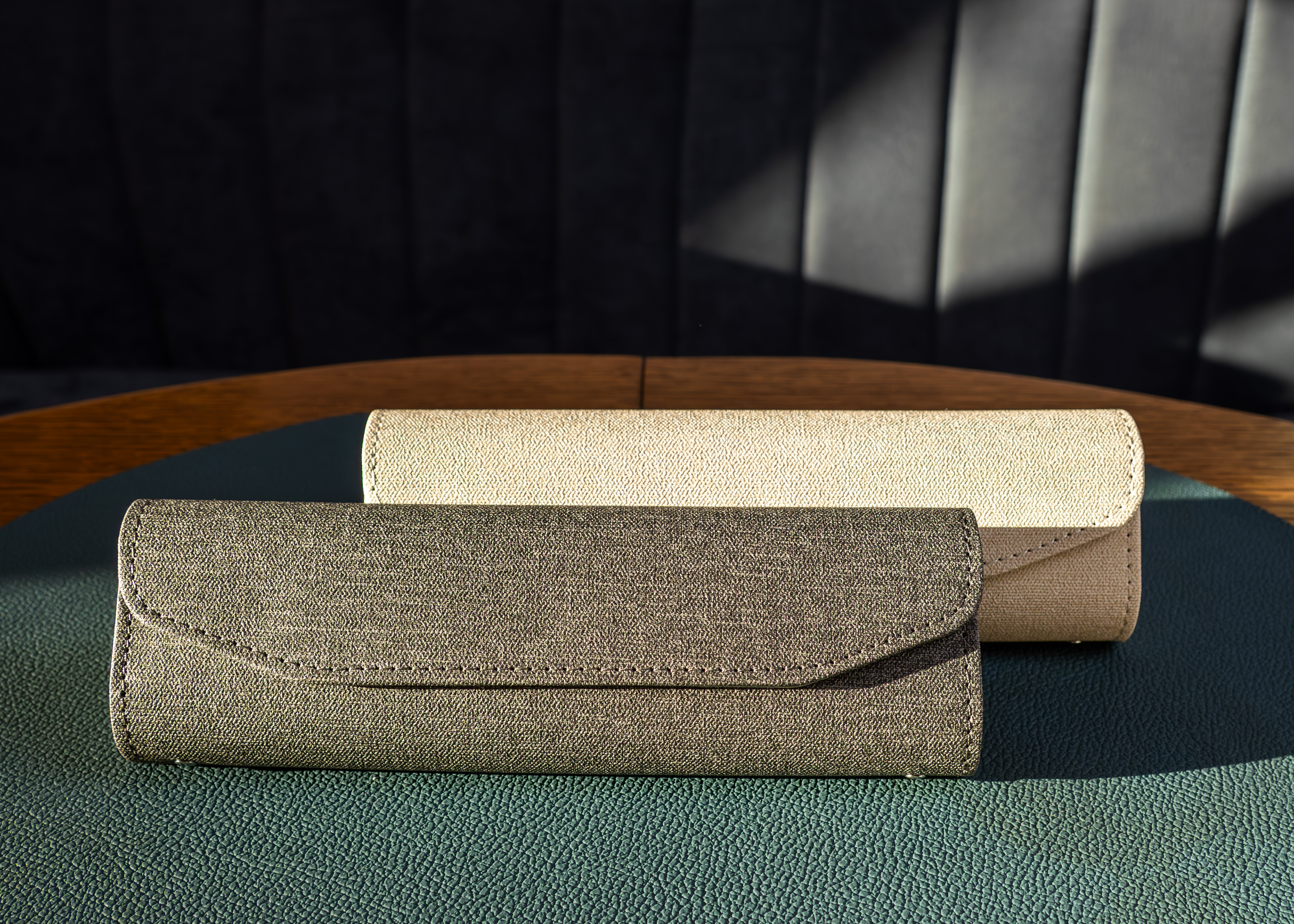 Dwa etui z tkaniny leżą na ciemnozielonej powierzchni. Jedno jest oliwkowe, drugie beżowe. Drewniany stół i ciemna ściana w tle.