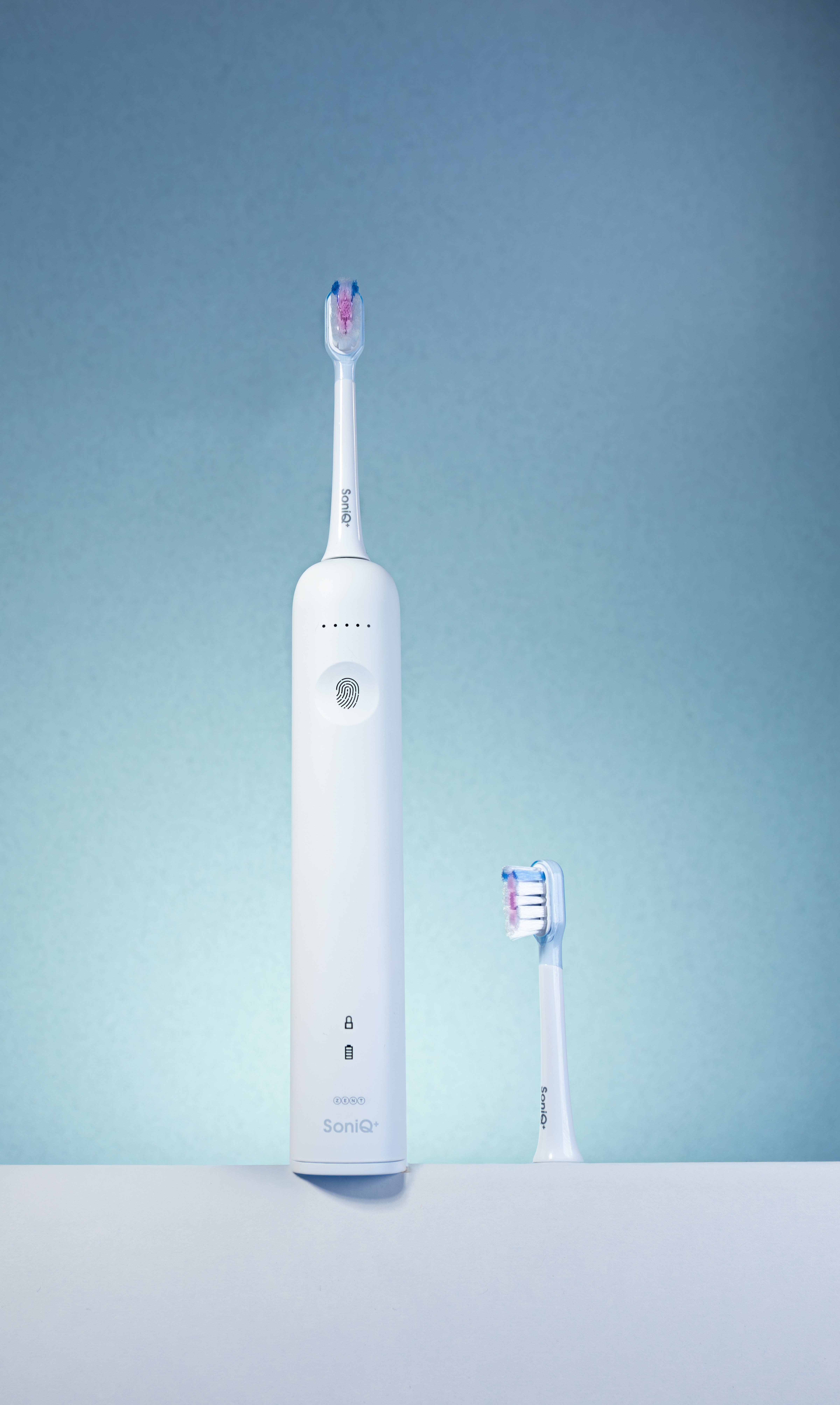 Biała elektryczna szczoteczka do zębów i główka szczoteczki stoją na białej powierzchni na niebieskim tle. Szczoteczka ma napis SoniQ+.