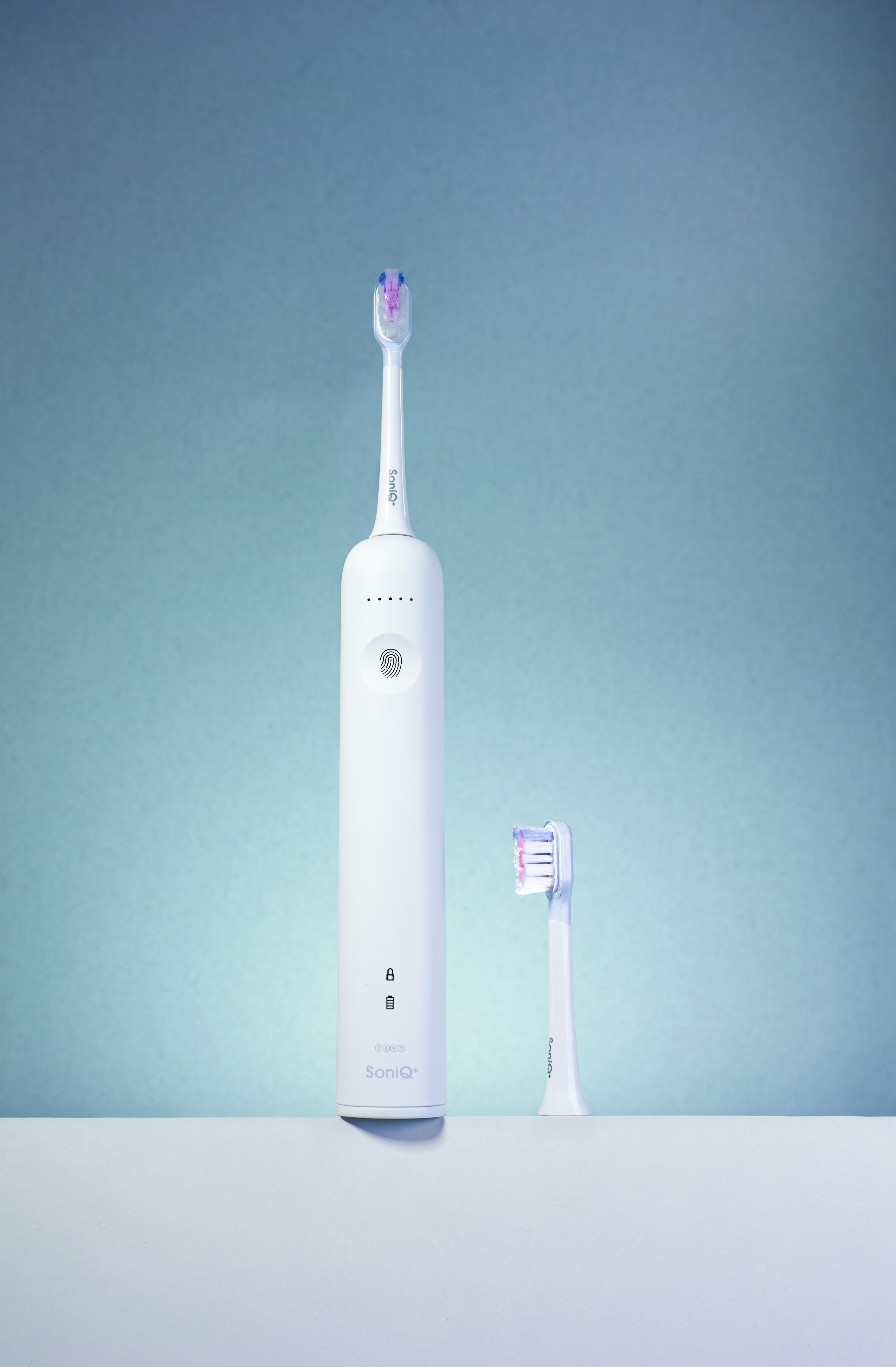 Biała elektryczna szczoteczka do zębów i zapasowa główka na białej powierzchni na niebiesko-zielonym tle. Szczoteczka ma napis SoniQ+.
