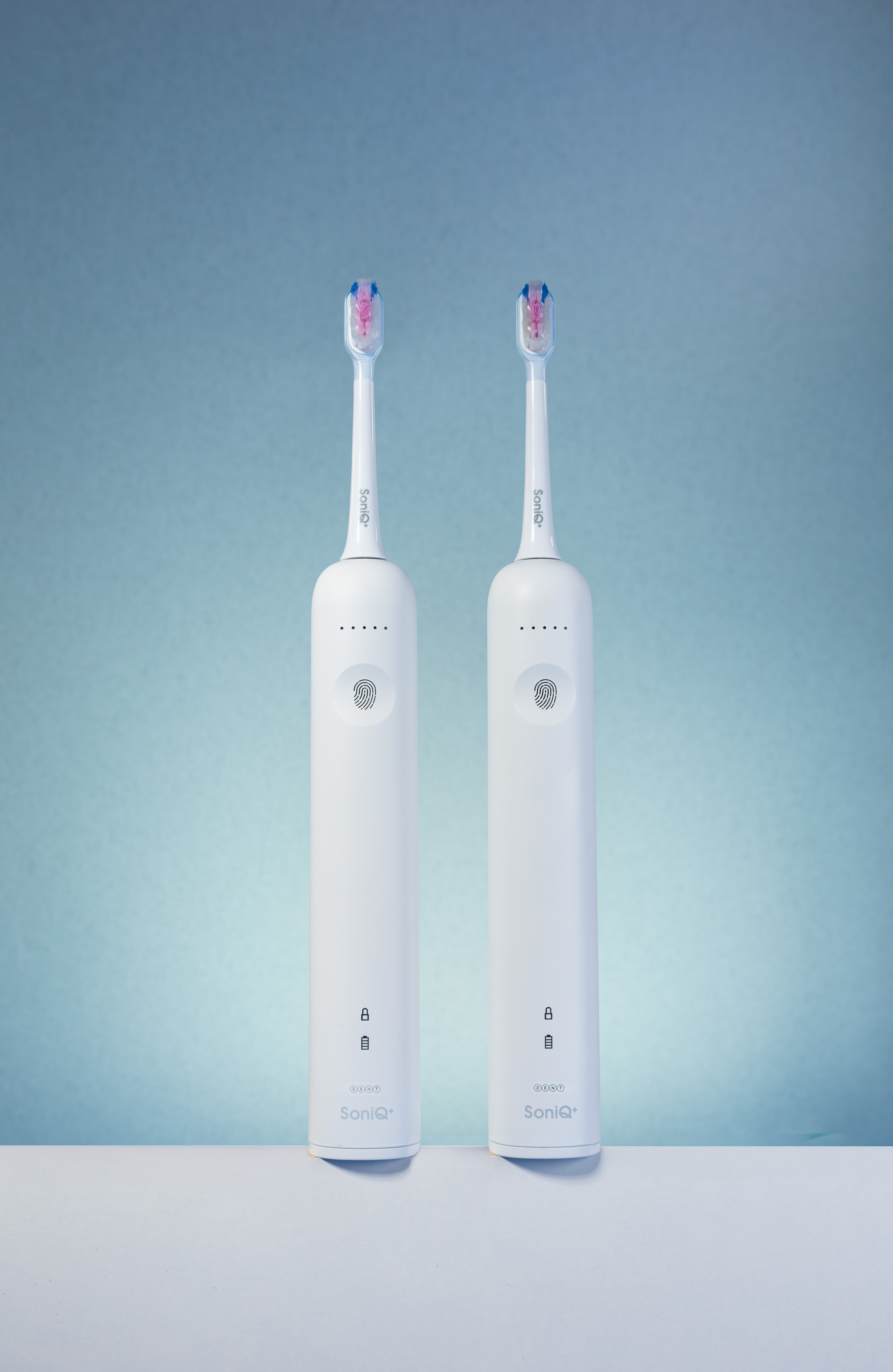 Dwie białe elektryczne szczoteczki do zębów z niebieskim i różowym włosiem na białej powierzchni, niebieskie tło. Marka: SoniQ+.