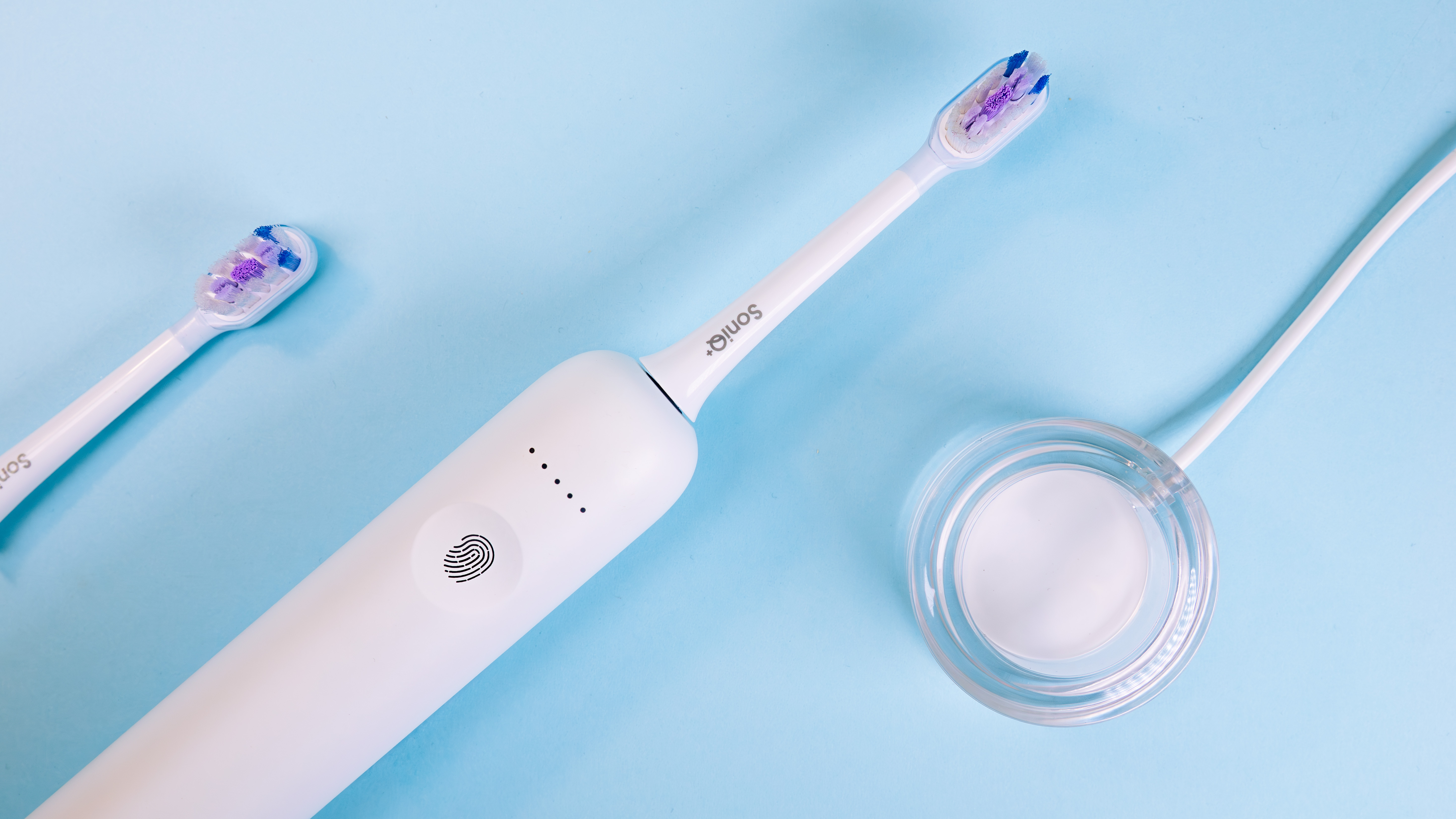 Biała elektryczna szczoteczka do zębów, zapasowa główka i stacja ładująca na jasnoniebieskiej powierzchni. Szczoteczka ma fioletowe włosie.