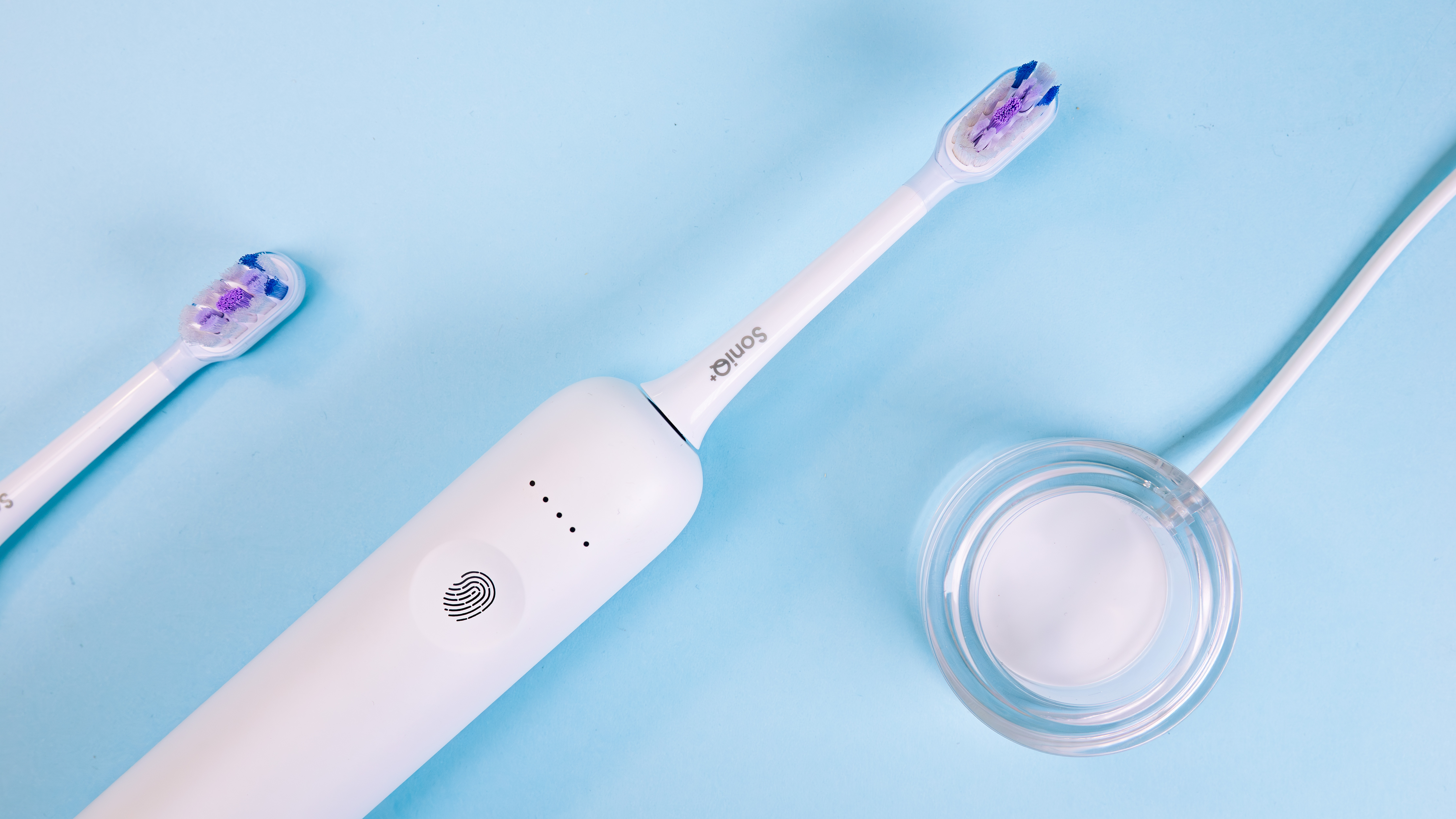 Biała elektryczna szczoteczka do zębów, stacja ładująca i zapasowa główka na niebieskim tle. Główki mają fioletowe i niebieskie włosie.
