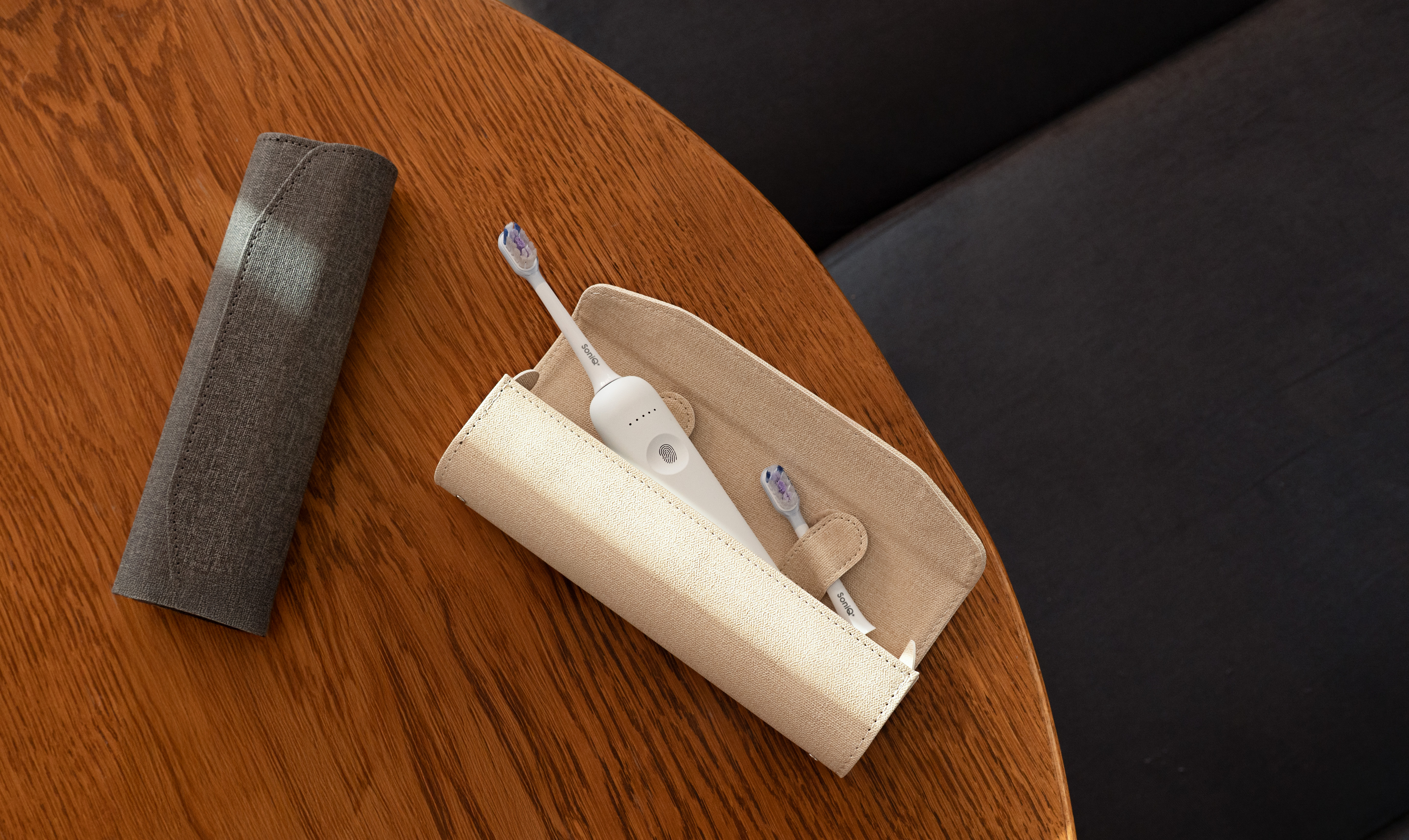 Biała elektryczna szczoteczka do zębów z dwiema główkami w beżowym etui na drewnianym stole. Obok szare etui.