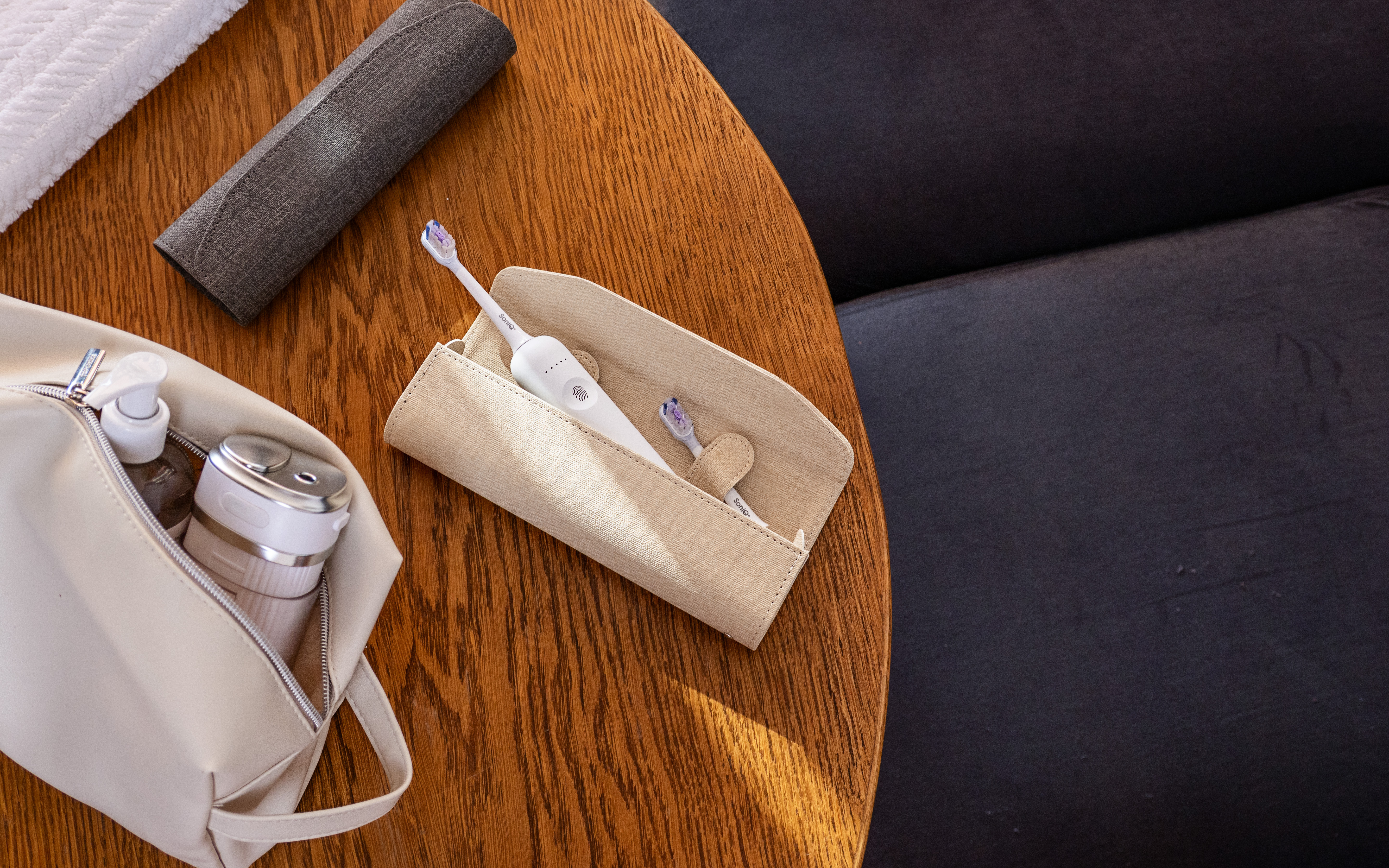 Na drewnianym stole etui na szczoteczkę, kosmetyczka, ręcznik i szara rolka. Po prawej stronie czarna powierzchnia.