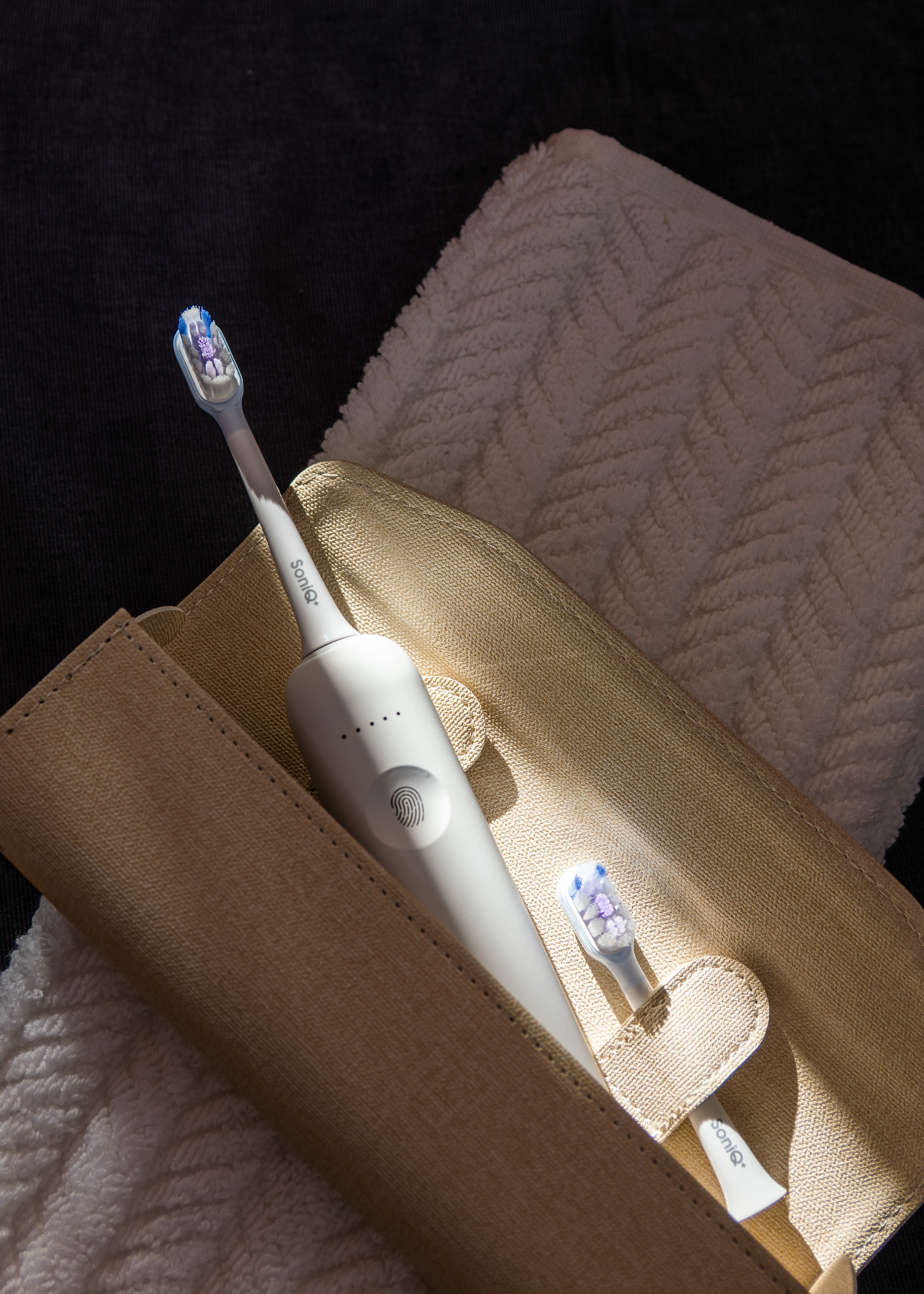 Szczoteczka elektryczna i zapasowa główka leżą w etui obok ręcznika, wszystko na ciemnej powierzchni.