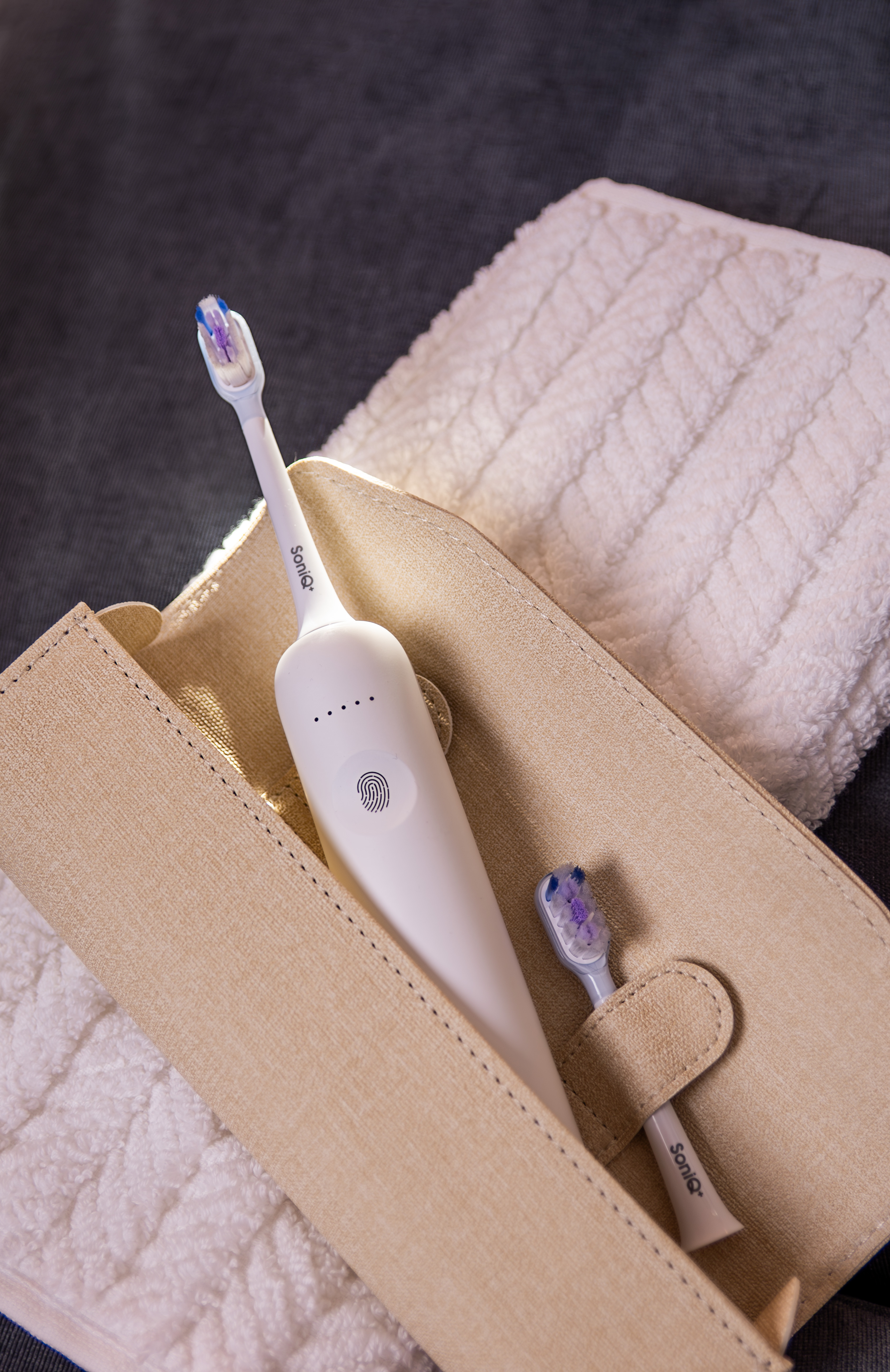 Biała elektryczna szczoteczka do zębów z dwiema główkami w beżowym etui, na białym ręczniku, wszystko na szarej powierzchni.