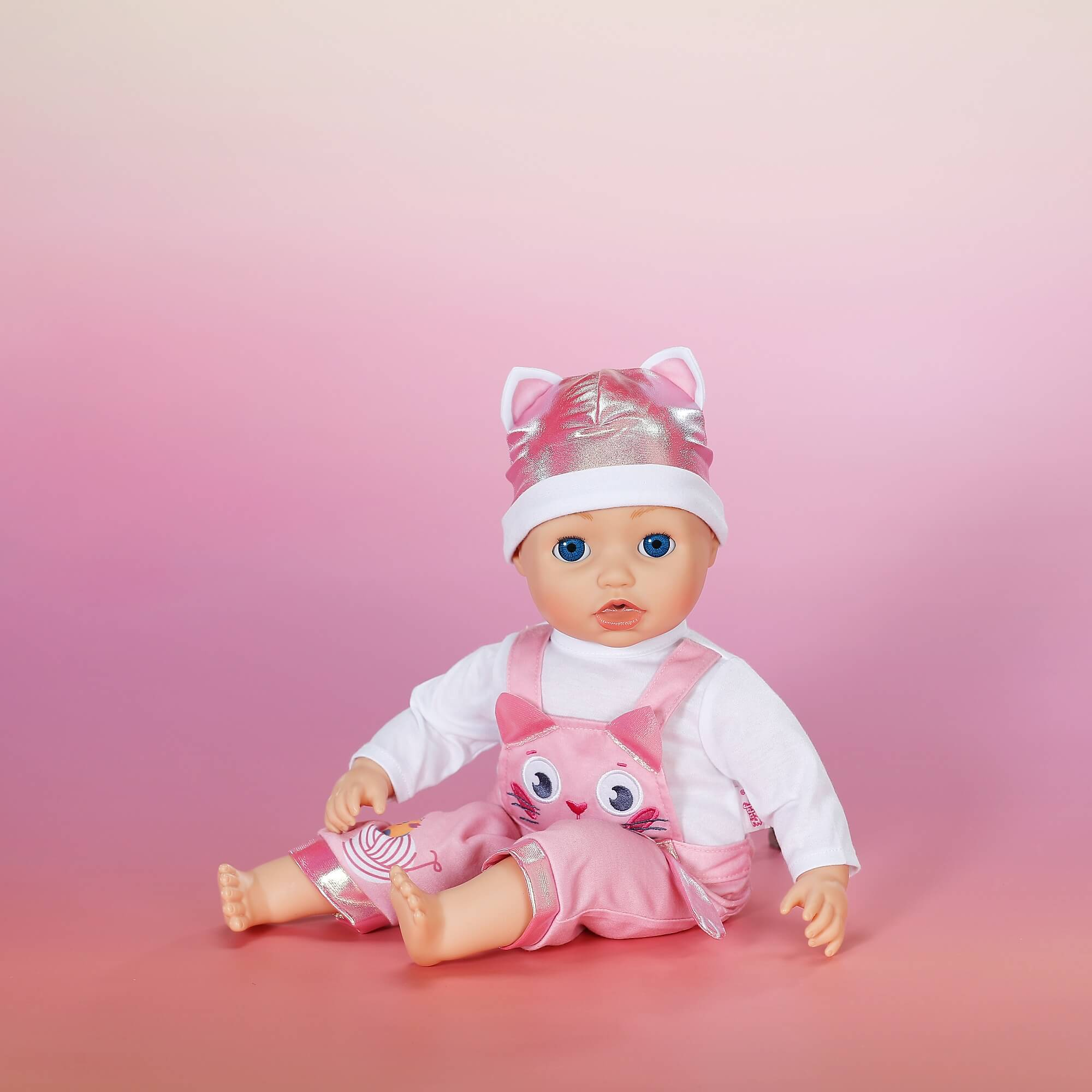 Lalka w różowej czapce i kombinezonie z kotem. Tło to różowy gradient.