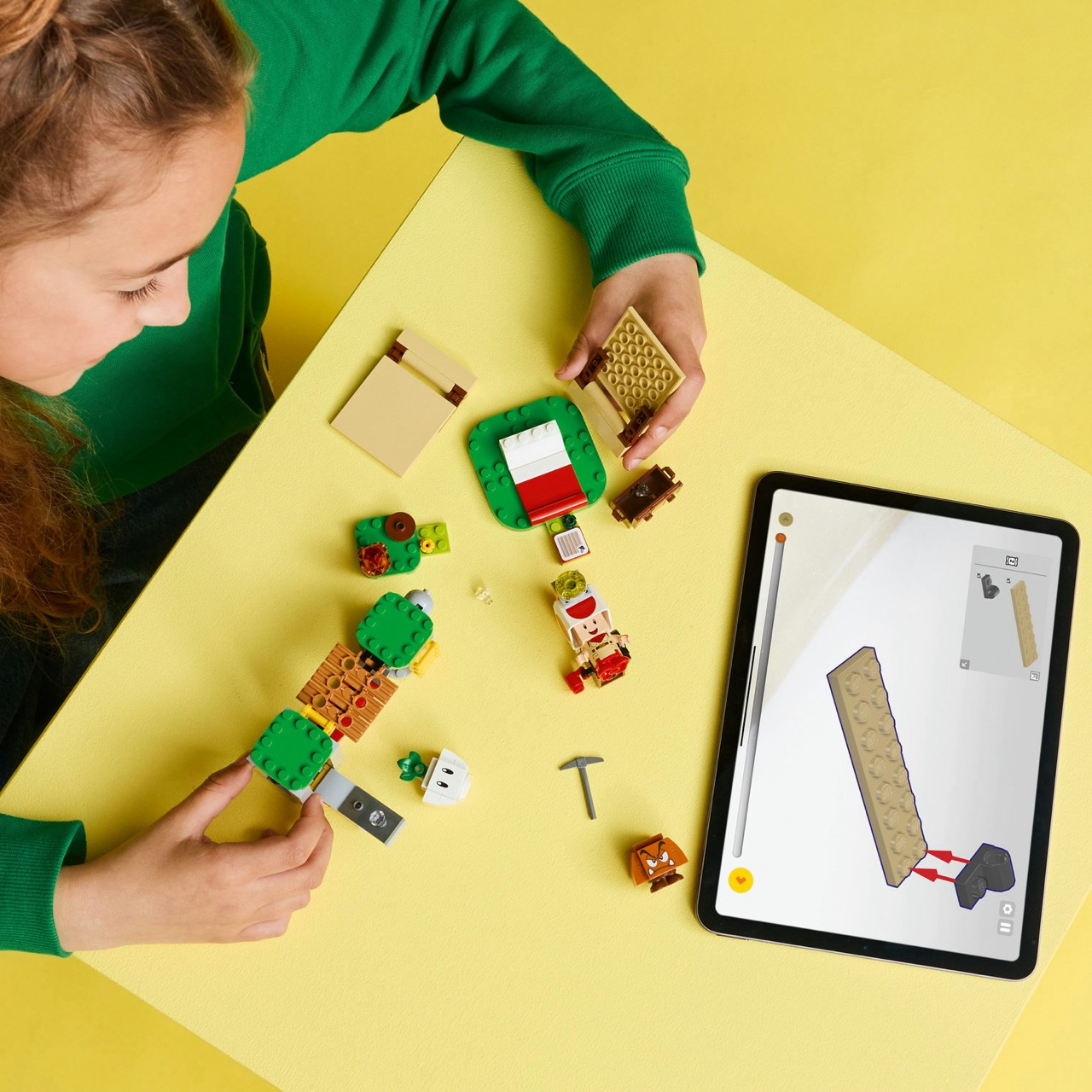 Una persona assembla pezzi Lego su una superficie gialla, guidata da un tablet. Varie figure e mattoncini Lego sono sparsi.