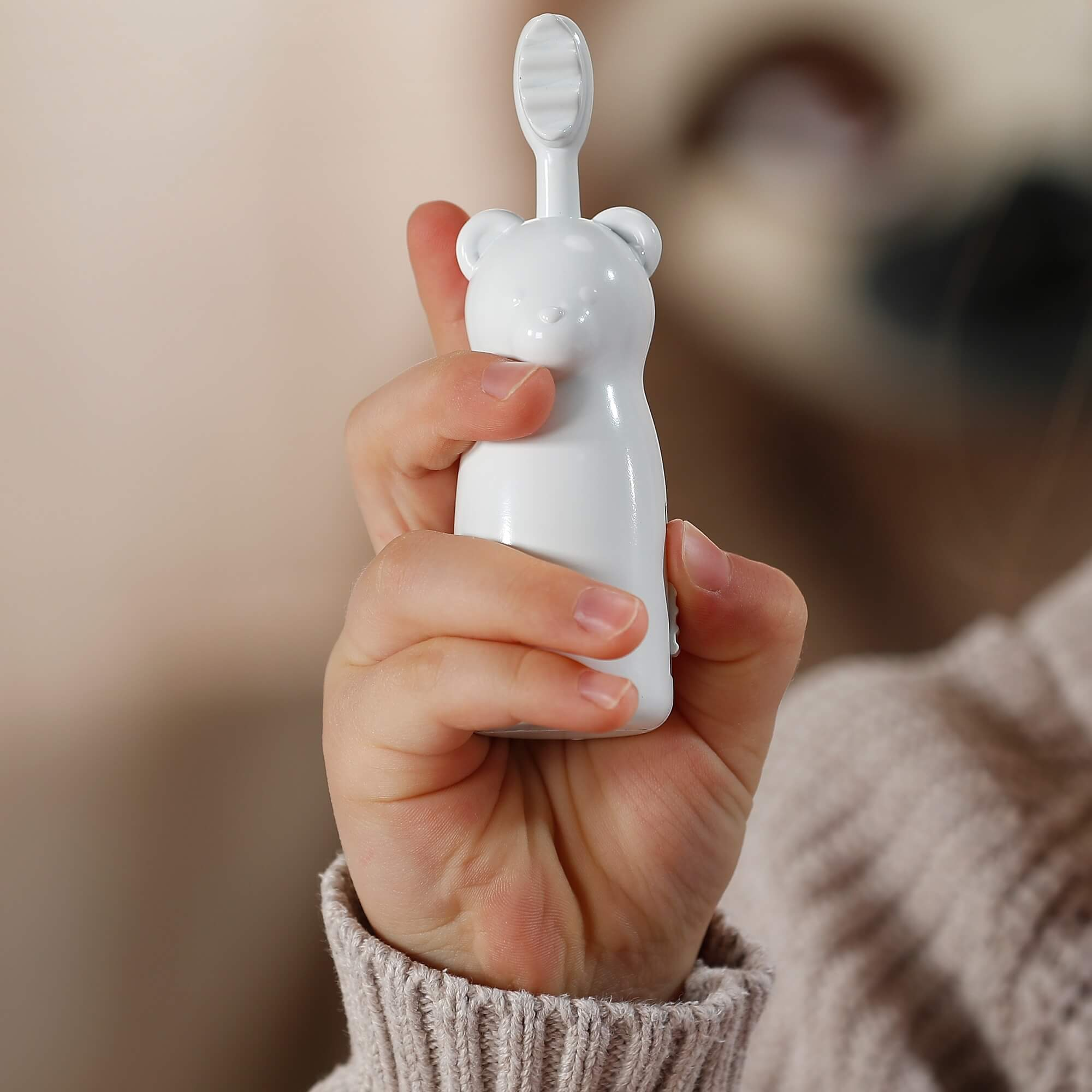 Ręka trzyma białą szczoteczkę do zębów w kształcie misia. Tło jest rozmyte, widać sweter i jasną ścianę.