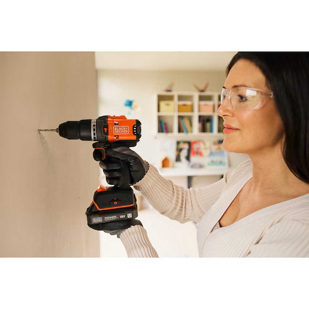 Bir kadın, BLACK+DECKER matkapla duvara delik açıyor. Güvenlik gözlüğü ve eldiven takıyor. Arka planda bir kitaplık var.