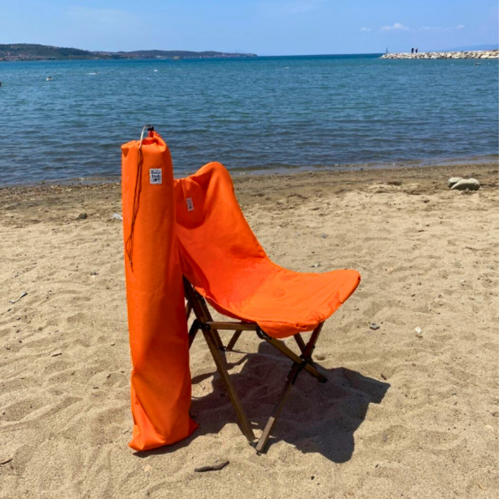 Turuncu bir plaj sandalyesi ve çantası kumlu bir sahilde duruyor. Arka planda deniz ve uzaktaki bir kara kütlesi var.