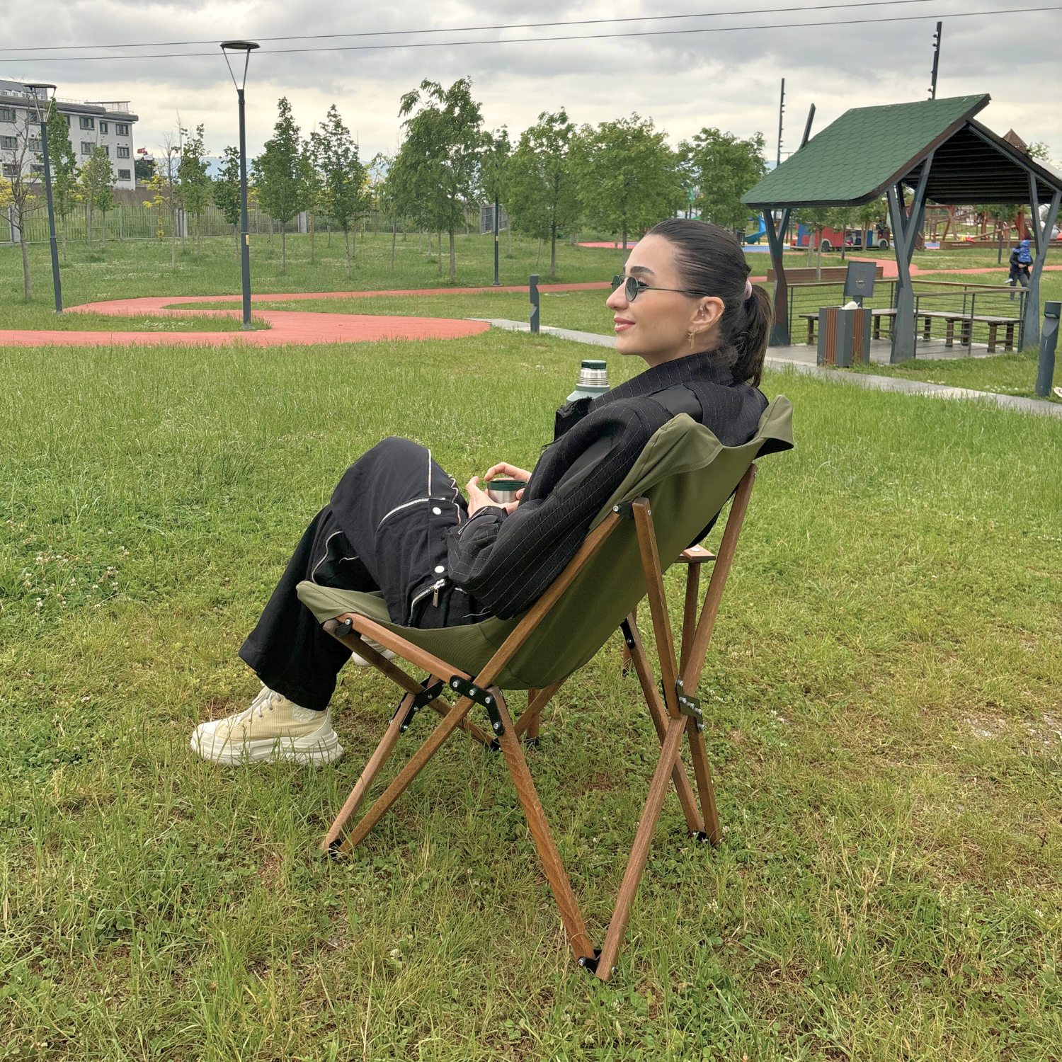 Bir kadın çimlerde bir sandalyede oturuyor, uzağa bakıyor. Güneş gözlüğü ve siyah giysiler giyiyor. Arka plan bir park.