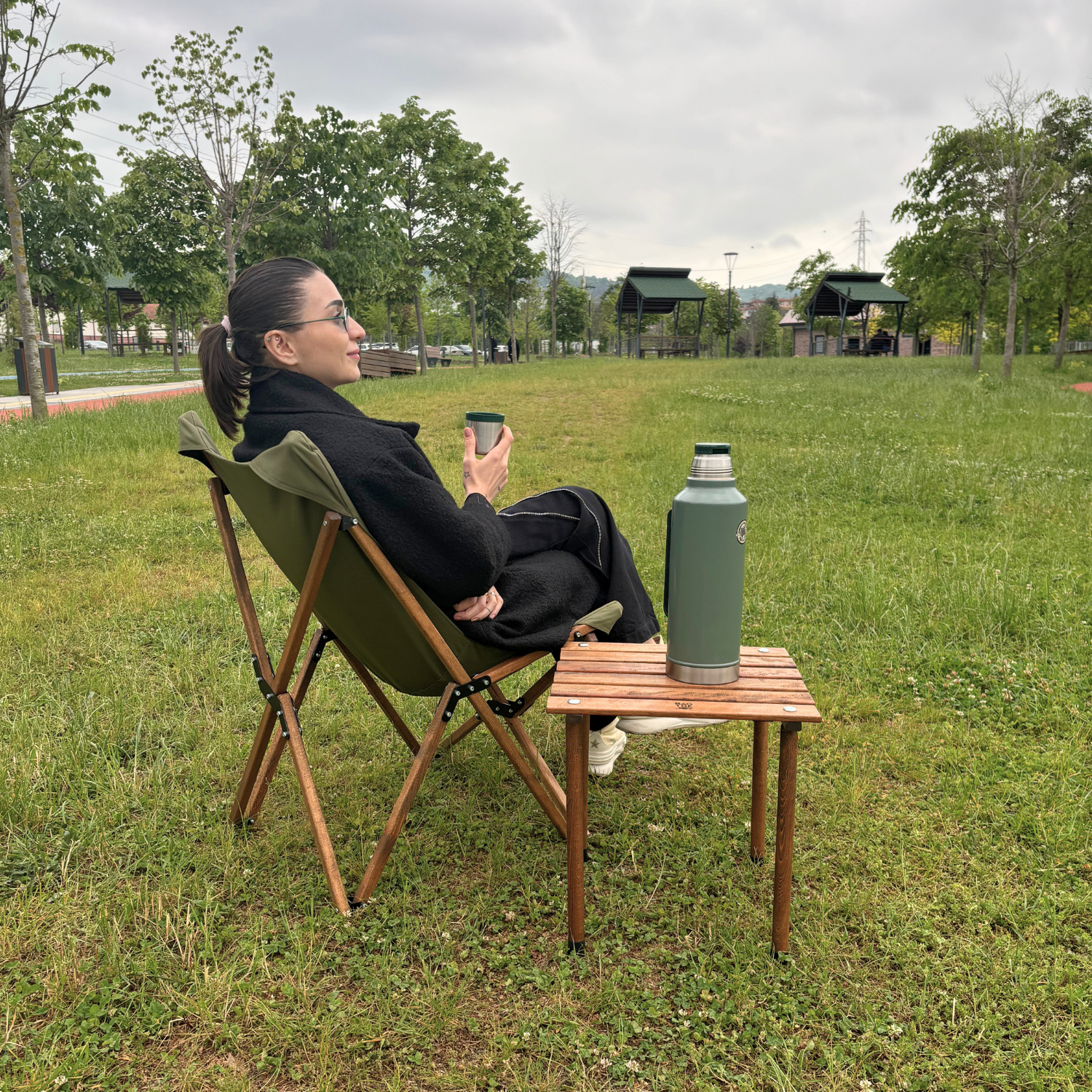 Siyah palto giymiş kadın park sandalyesinde oturuyor, fincan tutuyor. Yanında termoslu bir masa var. Arka planda yeşil çimen ve ağaçlar.