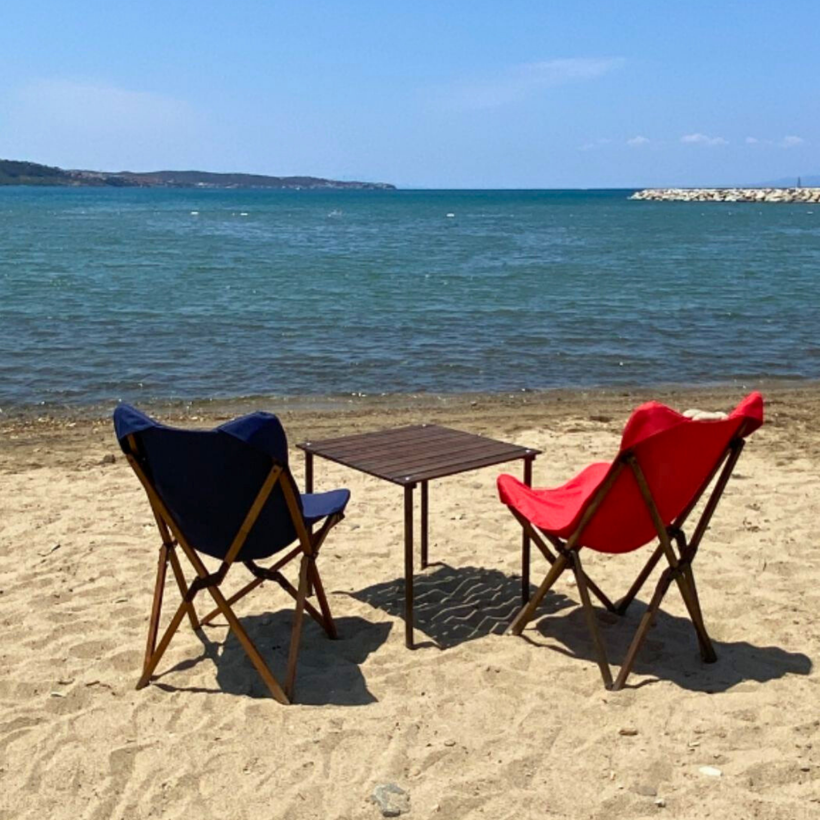 Mavi ve kırmızı sandalyeler, kumda küçük bir masa, arka planda deniz ve ada, açık gökyüzü ile plaj sahnesi.