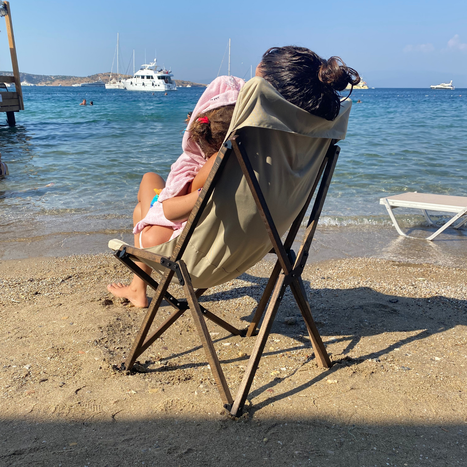 Bir kadın ve bir çocuk, kumlu bir plajda bir plaj sandalyesinde oturuyor. Arka planda yatlar ve deniz görülüyor.