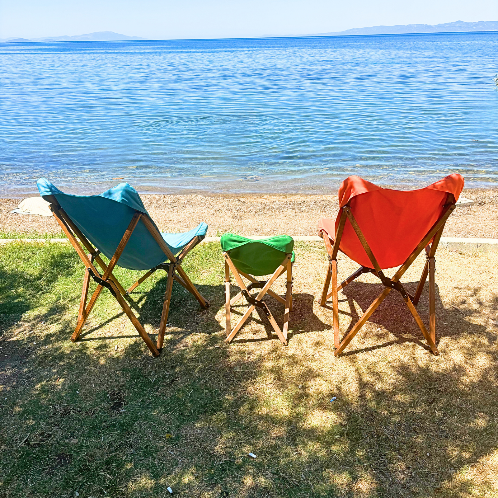 Mavi, yeşil ve turuncu üç katlanır sandalyeli plaj sahnesi. Arka planda deniz ve dağlar.