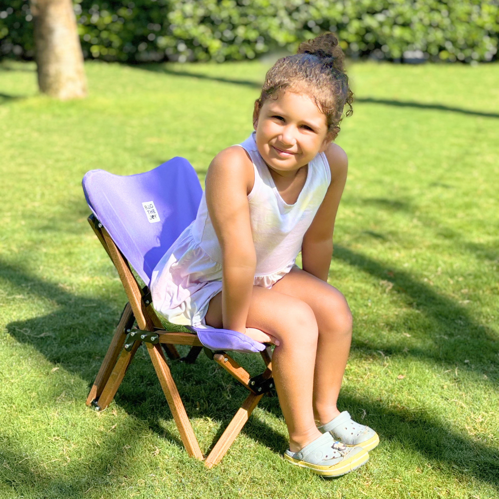 Güneşli bir parkta mor bir sandalyede oturan genç bir kız. Beyaz bir elbise giymiş ve kameraya gülümsüyor.